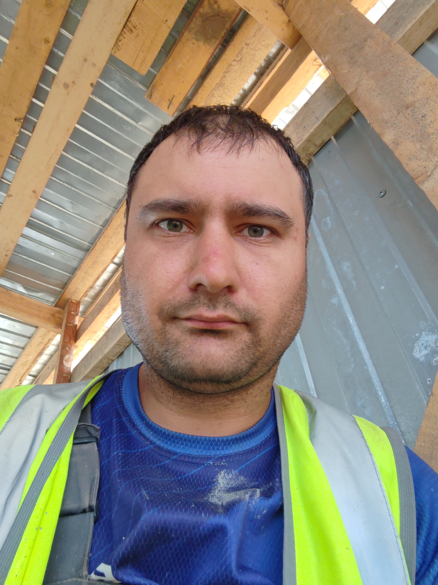 A medium shot selfie of a man wearing a blue shirt and a high-visibility vest is taken from a low angle outdoors. The man has short, dark hair, brown eyes, and stubble. He is looking directly at the camera with a neutral expression. The background consists of wooden beams and corrugated metal roofing, suggesting a construction site or unfinished building. The lighting is bright, casting some shadows on the man's face and the background. The overall impression is a candid snapshot taken in a work environment.