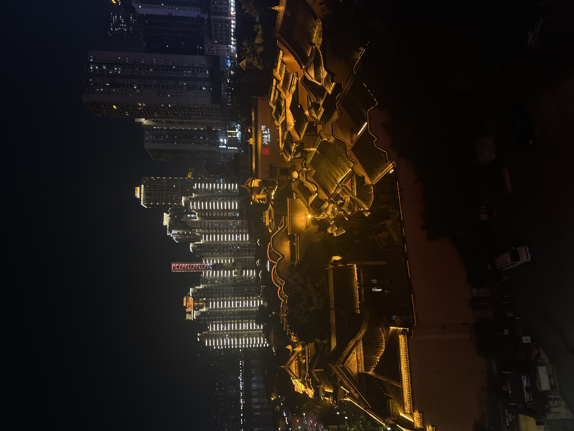 The image shows a night view of a city with a mix of traditional architecture and modern skyscrapers. The foreground features a complex of buildings with traditional Chinese-style roofs, illuminated by warm yellow lights along the eaves and outlines. These buildings appear to be part of a historical or cultural district. Behind this, a dense cluster of tall, modern apartment buildings rise into the night sky, their windows lit with white and yellow lights. In the far background, a body of water, likely a river, is visible, reflecting city lights. Some cars and streetlights are visible in the lower left portion of the image, indicating a street or road. The overall impression is of a vibrant, illuminated urban landscape at night, with a contrast between old and new architectural styles. The location is identified as Yuzhong District, China. There are no identifiable people in the image, and no specific actions or interactions are depicted.