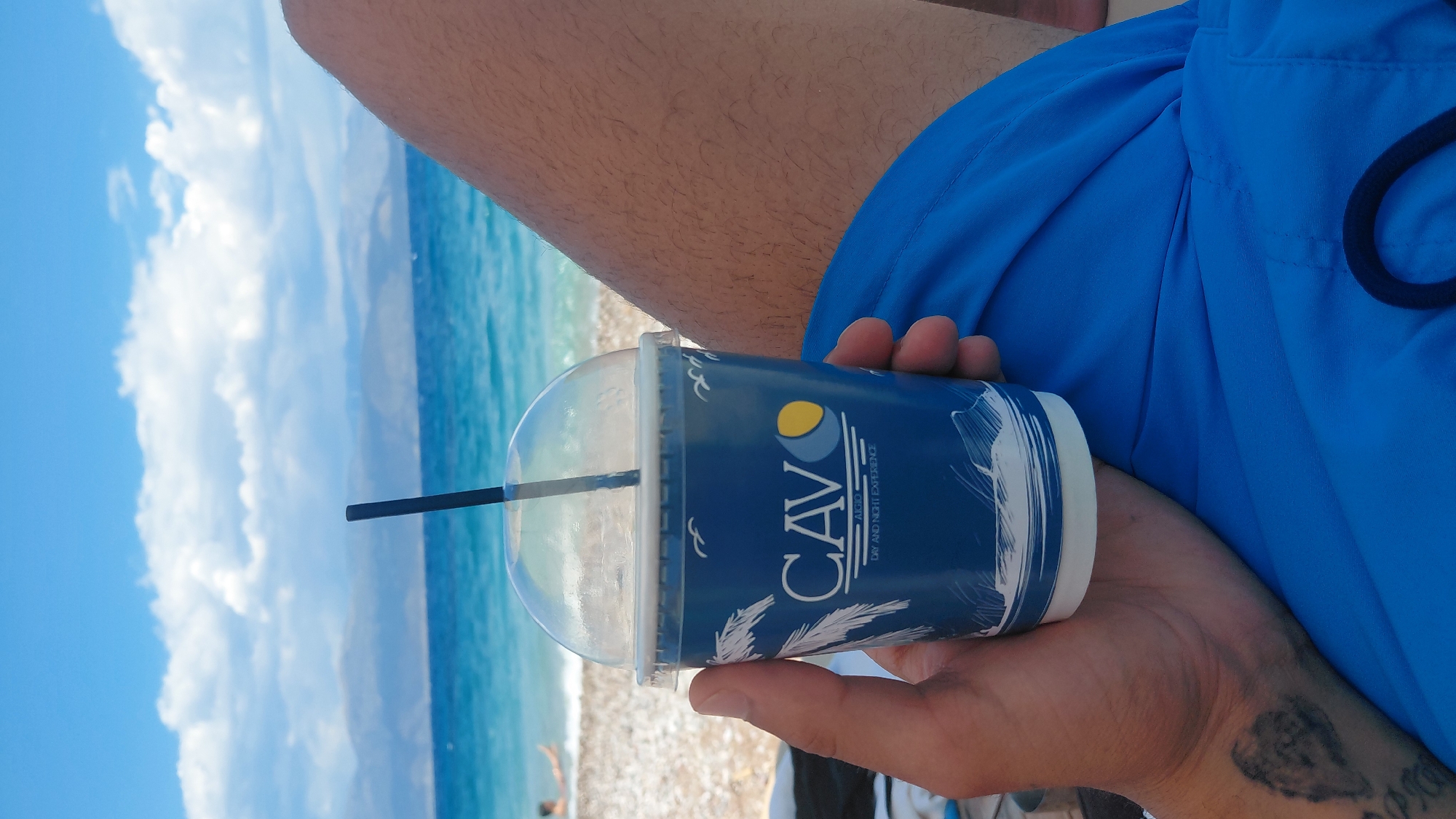 A close-up, eye-level, medium shot captures a person's hand holding a blue disposable cup with a clear plastic dome lid and a black straw. The cup is adorned with white palm fronds, seagulls, and the logo "CAVO AIGIO DAY AND NIGHT EXPERIENCE." The background reveals a sun-drenched beach with pebbles and the azure waters of the Gulf of Corinth, with hazy mountains in the distance under a sky dotted with fluffy white clouds. A person is visible in the shallow water on the left. The lower portion of the image is dominated by the person's thigh covered in blue swim trunks. A tattoo is visible on the person's forearm. The overall scene suggests a relaxing day at the beach in Aegio, Greece.