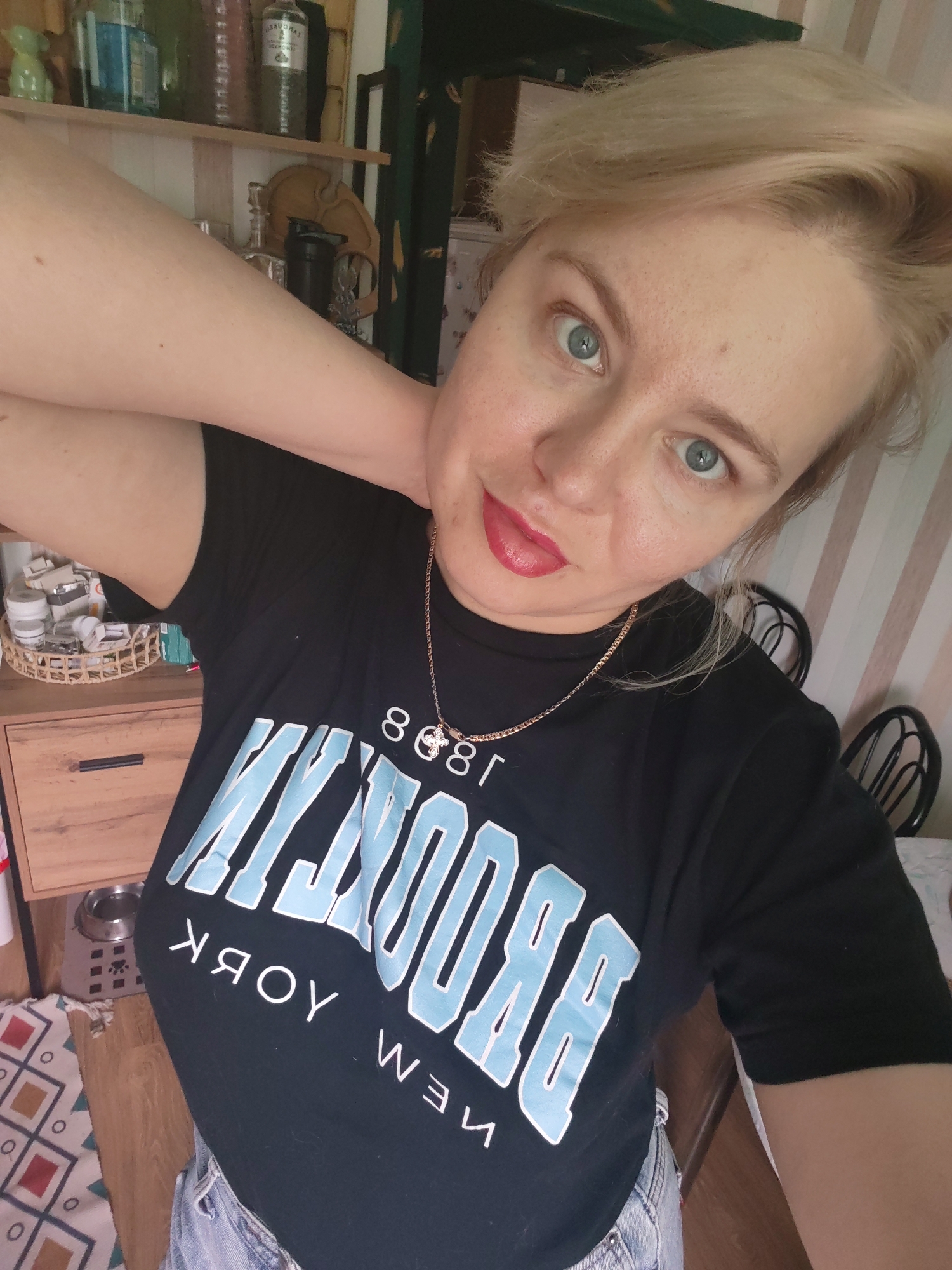 A woman with blonde hair and blue eyes is taking a selfie. She is wearing a black t-shirt with "Brooklyn New York" written on it in blue letters, and a gold chain with a cross pendant. The background appears to be a room with shelves filled with bottles and jars, and a wooden dresser. The flooring is a combination of wood and a patterned rug. The overall lighting suggests an indoor setting. The woman is resting her right arm on her neck. The setting is consistent with an interior space in Izhevsk, Russia, possibly a home.