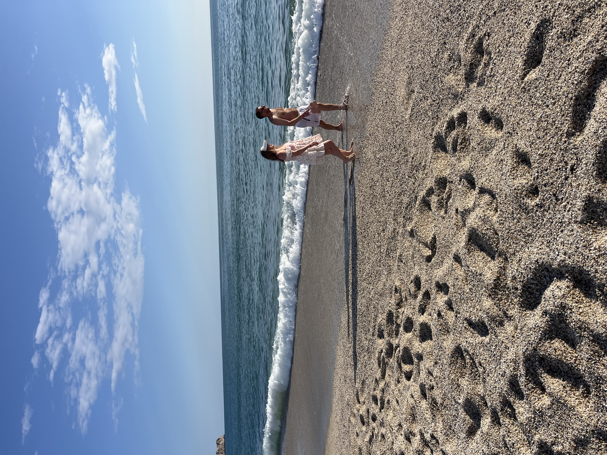 The image captures a sunny day at a beach in Alanya, Turkey. A man and a woman are standing near the water's edge. The man is wearing white swim trunks and sunglasses. The woman is wearing a light-colored dress with dark polka dots and a hat. The waves from the sea are breaking on the shore beside them. The beach sand is coarse and covered with footprints. Above, the sky is blue with scattered clouds.