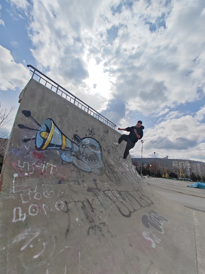 The image displays an outdoor concrete skatepark located in Shumen, Bulgaria, under a partly cloudy sky with direct sunlight breaking through. A single male individual, wearing a black hooded sweatshirt, dark trousers, and white athletic shoes, is captured mid-action on the inclined surface of a large concrete skate ramp. He is angled with his left foot firmly on the ramp and his right leg bent and lifted, suggesting a dynamic movement. His right arm is extended outwards, and his left hand holds a small dark object, possibly a mobile phone, near his ear. The concrete ramp is extensively covered with graffiti, including a prominent blue and yellow megaphone design with radiating lines, a light blue cartoon creature with teeth, and numerous smaller tags and scribbles in various colors. A dark metal railing runs along the top edge of the concrete wall that forms the upper boundary of the ramp. In the background, multi-story residential buildings characteristic of urban Shumen are visible, situated against a backdrop of distant hills and bare trees on the left. The ground surface of the skatepark, appearing to be concrete or asphalt, extends to the right of the ramp, with faint structures like streetlights and a blue tarp visible further in the distance.