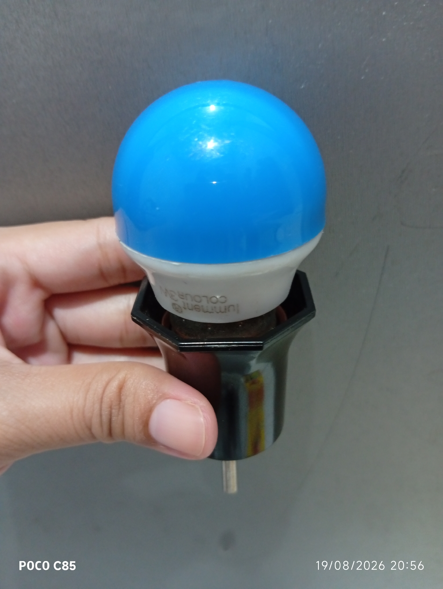 A close-up shot captures a hand holding a blue LED bulb with a black base. The bulb has white lettering on the base that reads "lumment @ colour 3W". The background is a plain gray textured wall. The timestamp "19/08/2026 20:56" and "POCO C85" are visible in the bottom right and left corners respectively, suggesting the image was taken in South Tangerang, Indonesia.