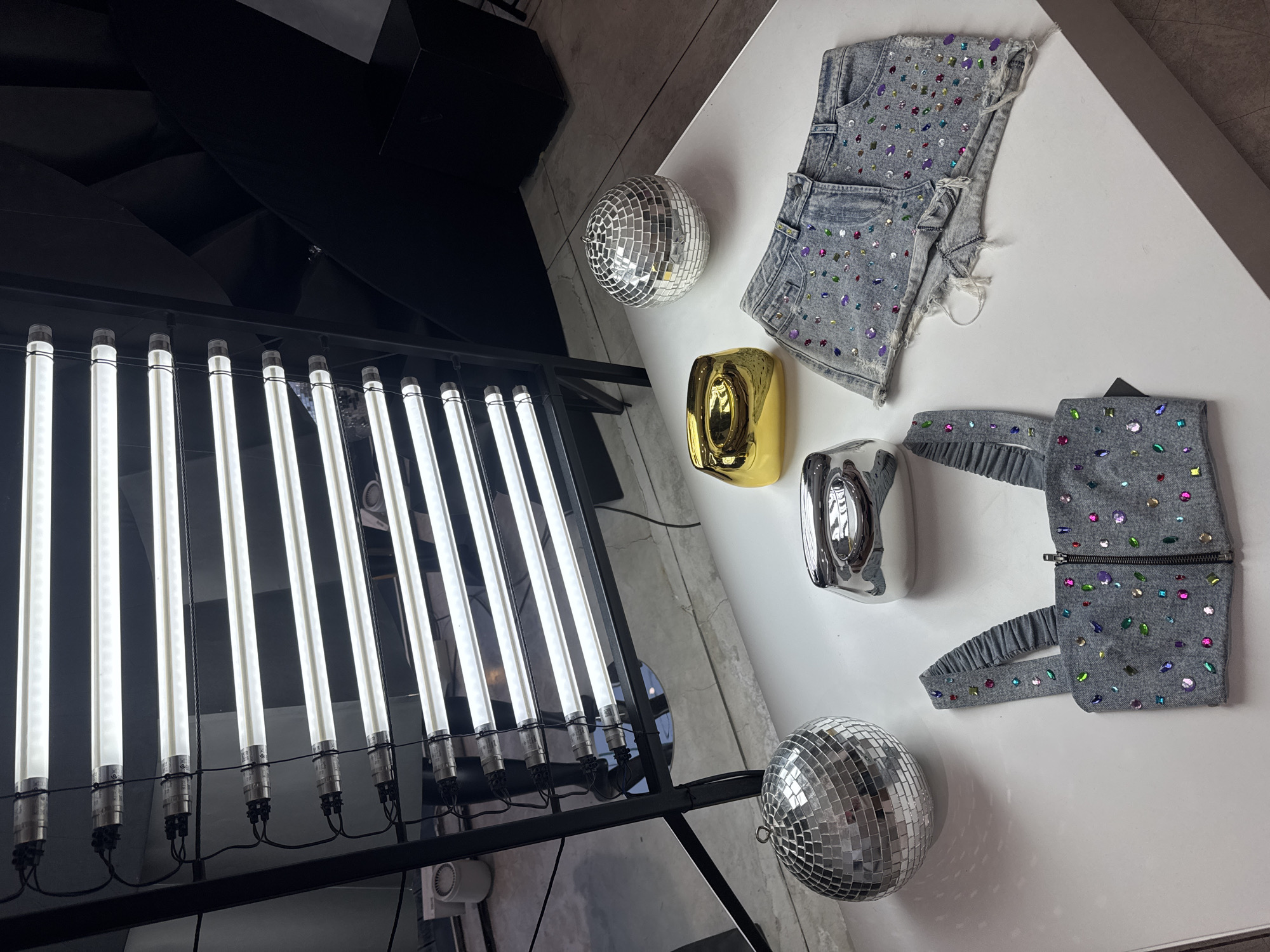 This image depicts a studio setup with multiple items displayed on a white table. In the background, a rack of linear fluorescent lights is positioned vertically, illuminating the scene. On the table, there are two disco balls, one to the left and another to the right. In front of the disco balls are two decorative bowls, one golden and the other silver. A denim crop top adorned with colorful rhinestones is laid out in the foreground, along with a pair of matching denim shorts also embellished with rhinestones. The overall environment suggests a fashion or product photography session. The location is stated to be North Kuta, Indonesia, though no specific landmarks or contextual clues within the image confirm this. There are no people visible in the image.