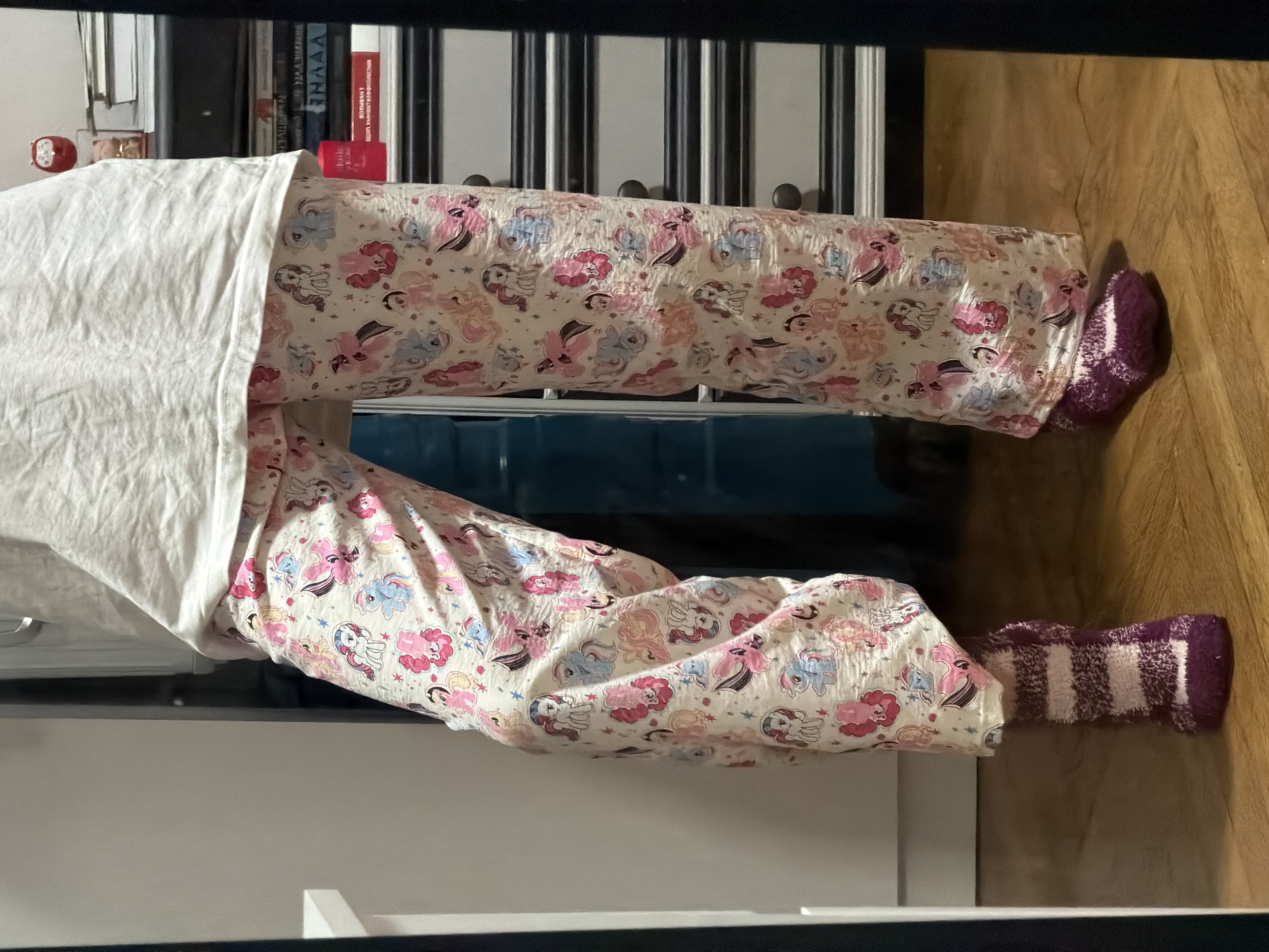 A person is standing in front of a mirror, wearing pajamas with a My Little Pony pattern and striped fuzzy socks. The pajamas are light-colored with various illustrations of My Little Pony characters in pink, blue, and white. The person is wearing one sock on their left foot, which has horizontal stripes of dark purple and white. The person's right foot is also wearing a similar striped sock. The background shows a closet or wardrobe with shelves filled with books and other items. The floor is made of wooden parquet. The overall setting appears to be a bedroom or dressing area. There is no specific indication of the location being in Moscow, Russia.