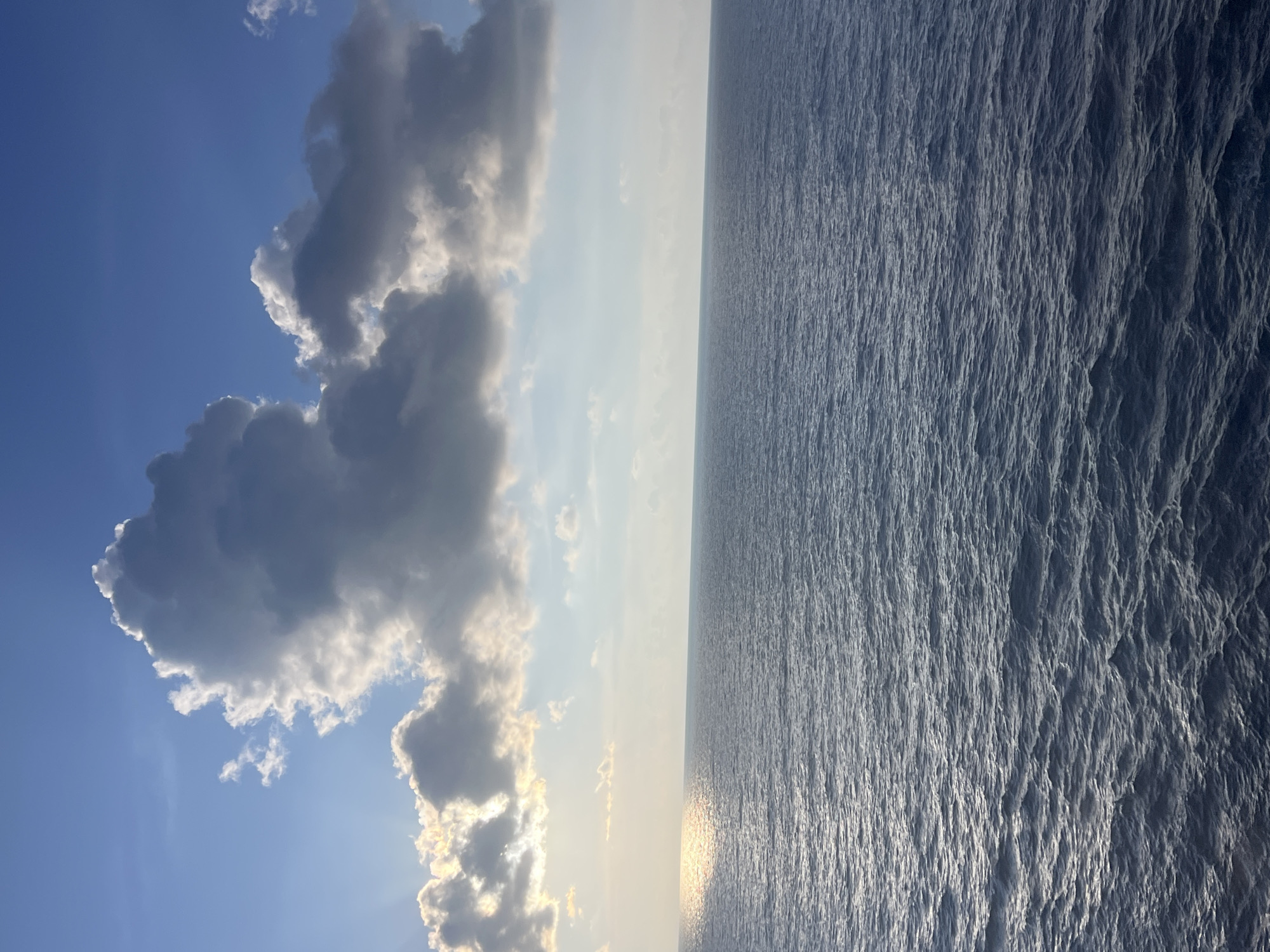 A wide shot of a body of water under a blue sky with clouds. The sun reflects off the water on the left side of the image, casting a bright light. A large, dark cloud is prominent in the sky, with smaller clouds scattered around it. The water has small waves.