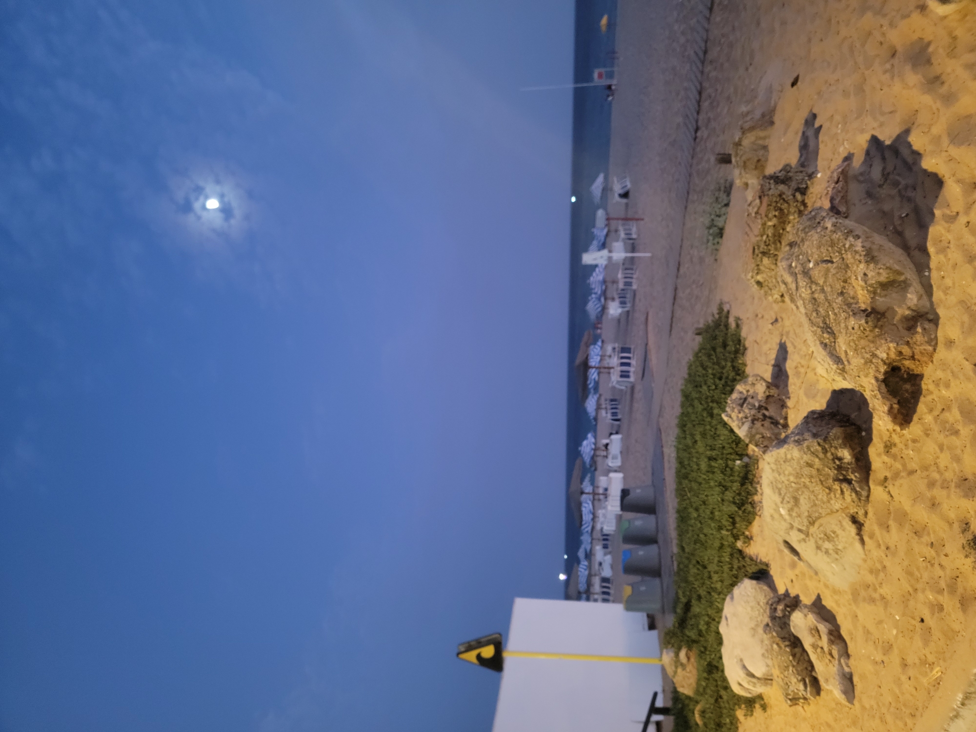 The image depicts a beach scene in Quarteira, Portugal, at twilight. The sky is a deep blue with scattered clouds, and the moon is visible in the upper right quadrant. In the foreground, several large rocks are scattered on the sandy beach. Behind the rocks, there is a patch of green vegetation. Further back, a row of white beach umbrellas with blue and white striped canopies are set up, along with white lounge chairs. A white building with a yellow pole and a triangular warning sign is visible on the left side of the image, with several trash bins next to it. The ocean stretches across the horizon, with a faint light visible on the water in the distance. The scene appears calm and empty, suggesting it is either early evening or late night.