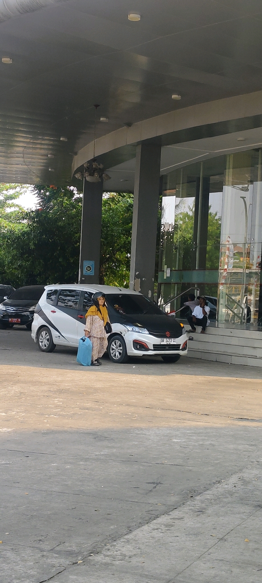 A woman in traditional clothing, including a headscarf and patterned suit, stands next to a white and black Suzuki Ertiga car. She is holding a large blue plastic bag in her right hand. The car has a license plate that reads "DA 1473 CE". Behind her, to the right, a man in a white shirt and dark pants sits on the steps of a building. The building has large glass windows and a gray, modern architectural style. Trees with green foliage are visible in the background, suggesting an outdoor setting. The ground is paved with concrete. The scene appears to be in a public area, possibly a hotel or commercial building entrance in Banjarmasin, Indonesia, as indicated by the license plate prefix "DA" which is used in Kalimantan.