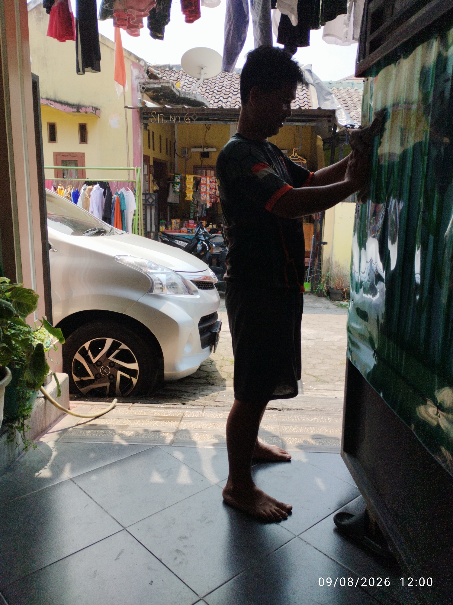 A man is cleaning a green, corrugated plastic surface. He is wearing a dark t-shirt with orange trim on the collar and sleeves, and dark shorts. He is barefoot. Beside him is a white car, and beyond that, a clothesline with laundry hanging. In the background, there is a building with a red-tiled roof and yellow walls, and a satellite dish. The scene appears to be outdoors, possibly a residential area. The timestamp "09/08/2026 12:00" is visible in the lower right corner.