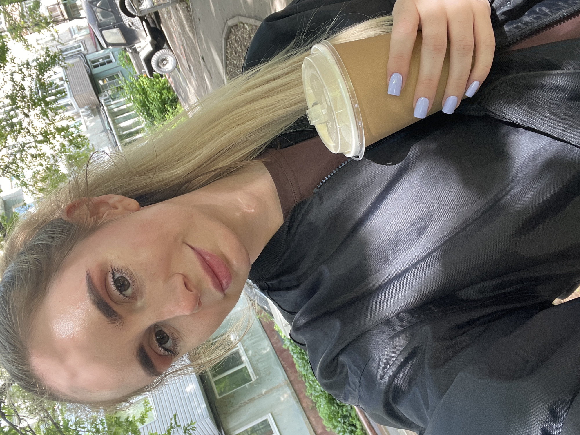A selfie captures a woman with blonde hair holding a coffee cup in an outdoor setting. She wears a brown top under a black jacket. Her fingernails are painted a light purple. The background features buildings and foliage, typical of an urban environment. In the distance, a vehicle can be seen. The setting appears to be a street in a populated area. The image does not provide sufficient information to pinpoint the exact location as Vladivostok, Russia.