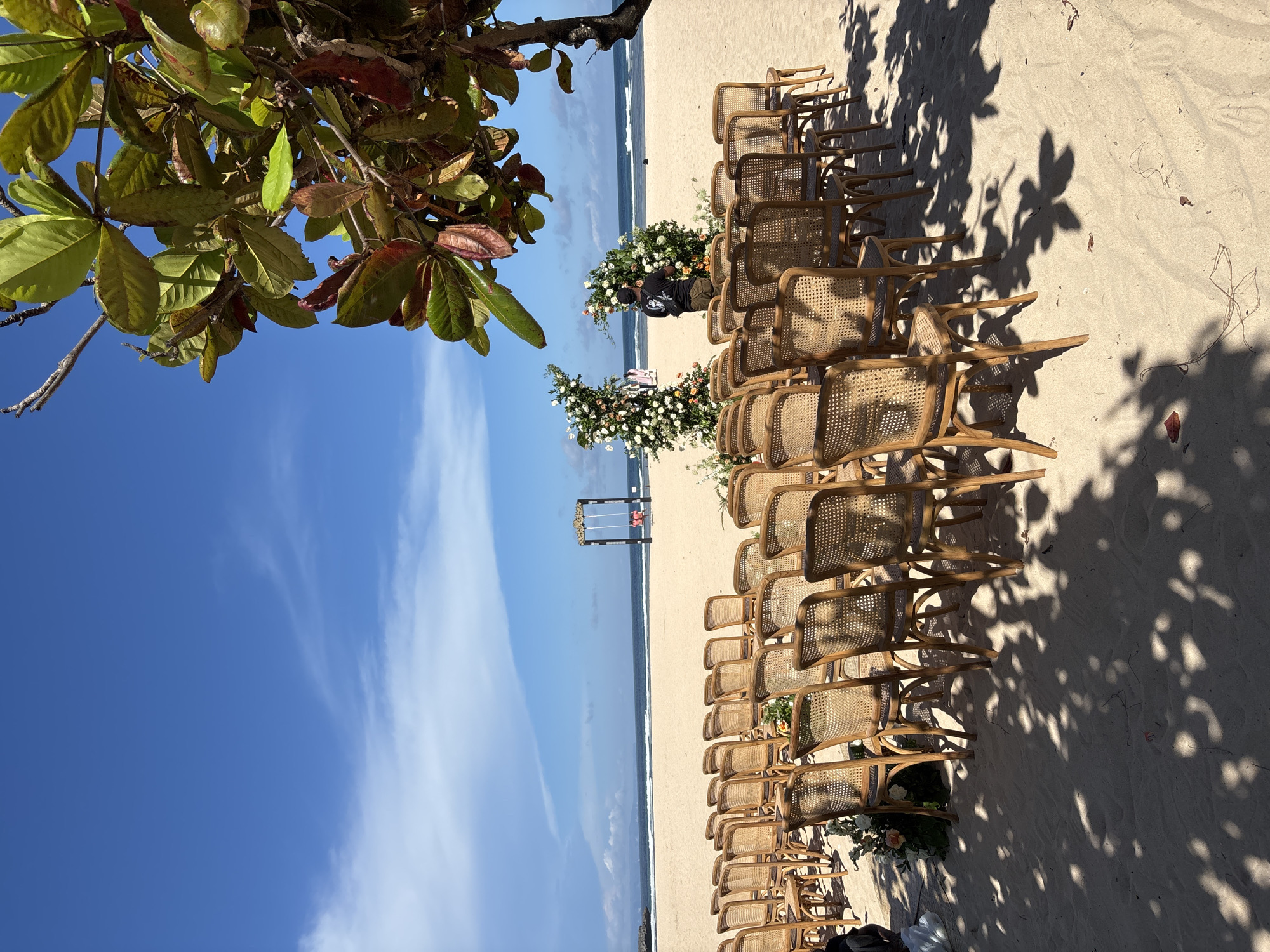 The image captures an outdoor event setup on a sandy beach, likely a wedding, in Nusa Dua, Indonesia. Rows of wooden chairs are arranged on either side of a central aisle leading to a decorated wedding arch in the distance. Floral arrangements adorn the sides of the aisle. In the background, the ocean meets the sky. A person can be seen amongst the floral arrangements. The sky is blue with some clouds. A tree with green and brown leaves overhangs the scene from the top left. Shadows from the tree are cast on the sand around the chairs.