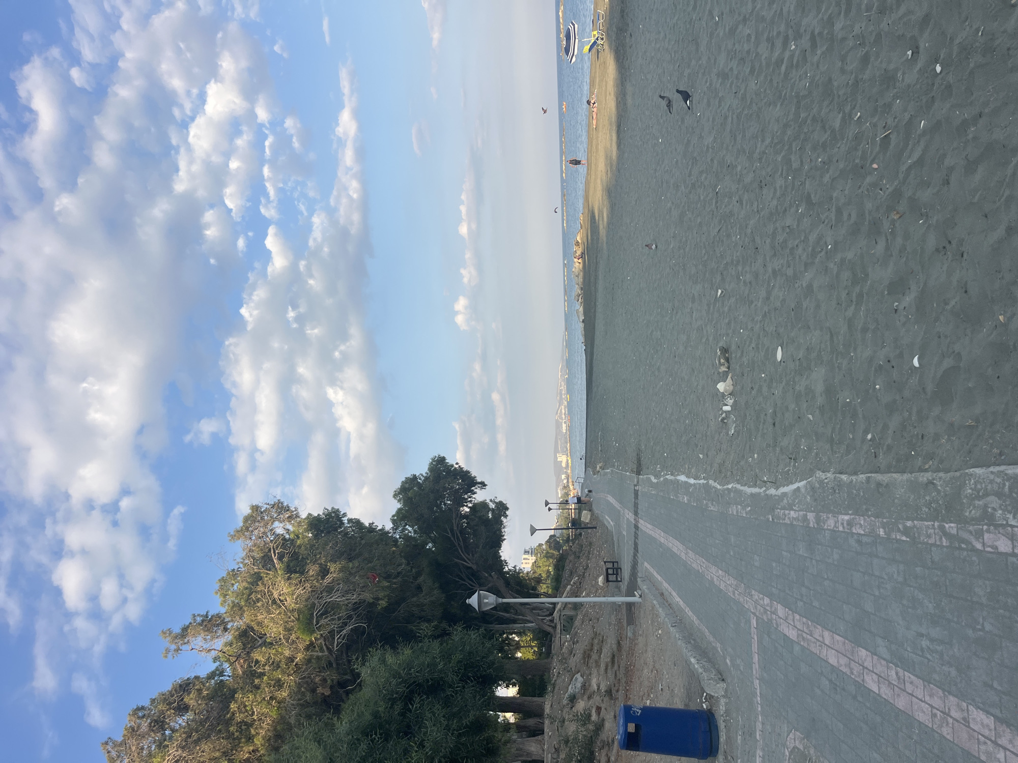 This is an eye-level, medium shot of a coastal scene in Agios Tychona Community, Cyprus, during the day. The foreground is dominated by a dark sand beach. To the left, a paved walkway with pink accents runs alongside the beach, bordered by trees on the far left and a blue trash bin. Further down the walkway, a person is walking a dog. In the mid-ground, people are visible on the beach, some sunbathing and one standing near the water. Several boats are scattered across the calm sea. The background shows a distant city skyline. The sky is a light blue with scattered white clouds, and the overall lighting suggests it is late afternoon.