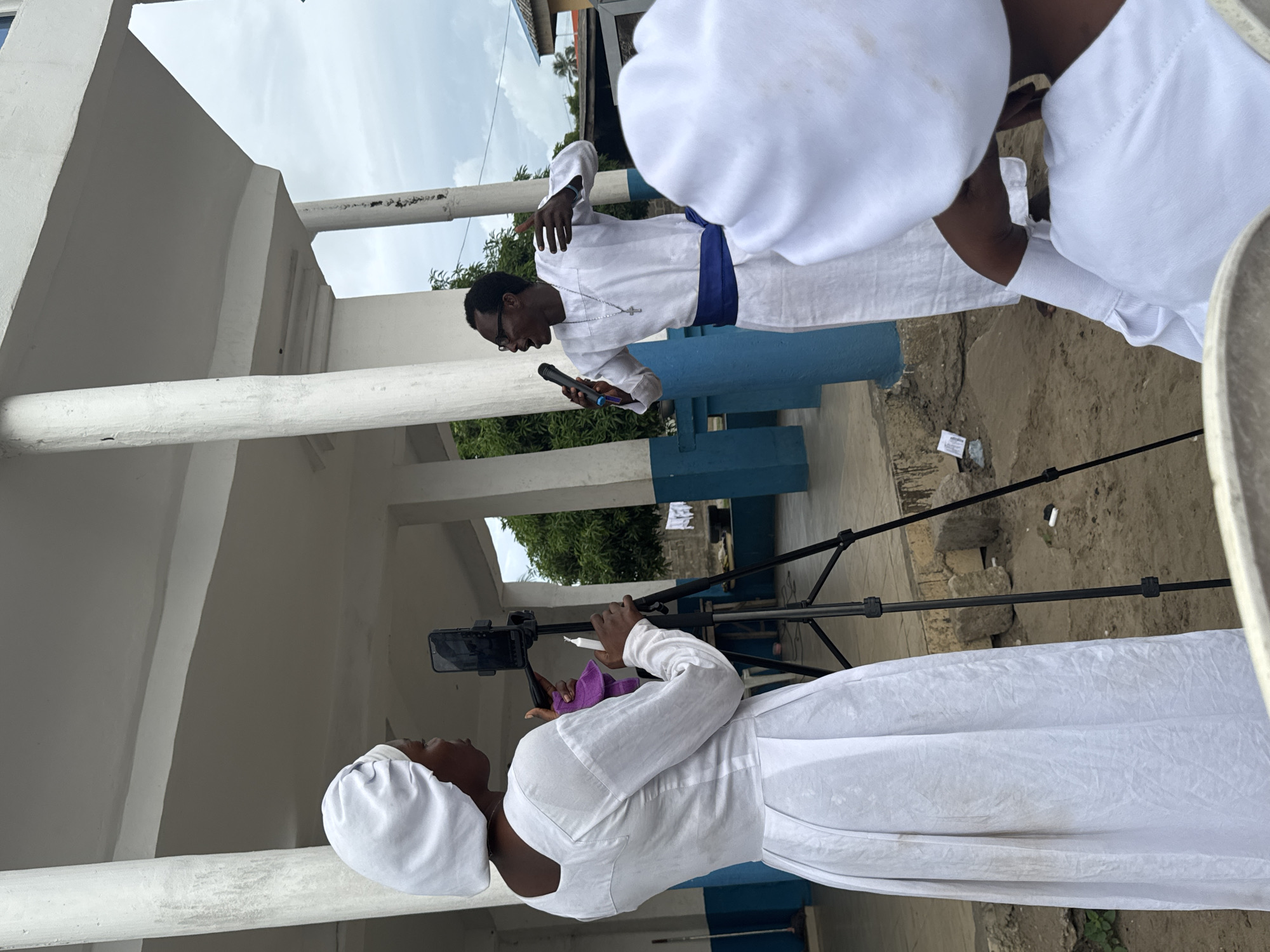 The image depicts a scene in Badagry, Nigeria, featuring people in white attire. A man in a white robe with a blue sash is speaking into a microphone. Another person is recording him with a smartphone mounted on a tripod. Other people wearing white garments and hats are visible in the background. The setting appears to be outdoors, adjacent to a building with white columns and blue accents. Debris is scattered on the ground. The sky is overcast.