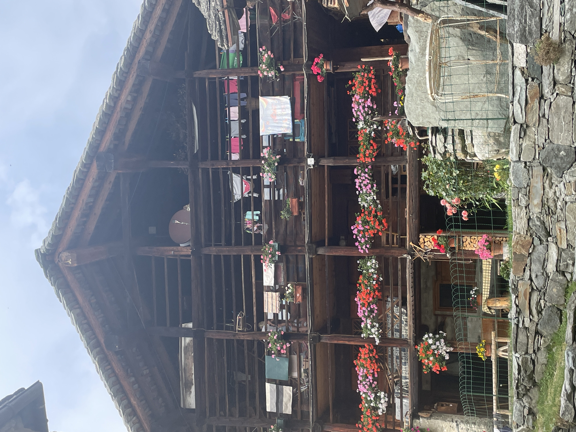 The image depicts the exterior of a traditional wooden chalet in Alagna Valsesia, Italy, against a backdrop of a cloudy sky. The chalet features multiple levels of wooden balconies adorned with numerous potted plants and hanging flower baskets. On the balconies, various items are hung, including laundry and what appear to be textiles or rugs. A person is partially visible on one of the upper balconies. The foreground is characterized by a stone wall, grass, and a green wire fence enclosing a small area with a metal table and chairs. The architectural style suggests a rustic, possibly historical, mountain dwelling.