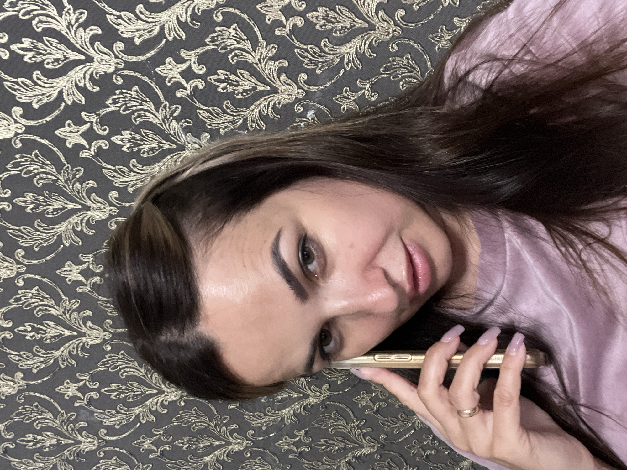 A woman is holding a gold-colored smartphone to her right ear, her left hand resting near her cheek. She is looking towards the camera with a neutral expression. The background is a dark gray wallpaper with a repeating gold damask pattern. The woman has long, dark brown hair and is wearing a pinkish-purple, silky top. A gold ring is visible on her left ring finger. The image appears to be a selfie, taken indoors.