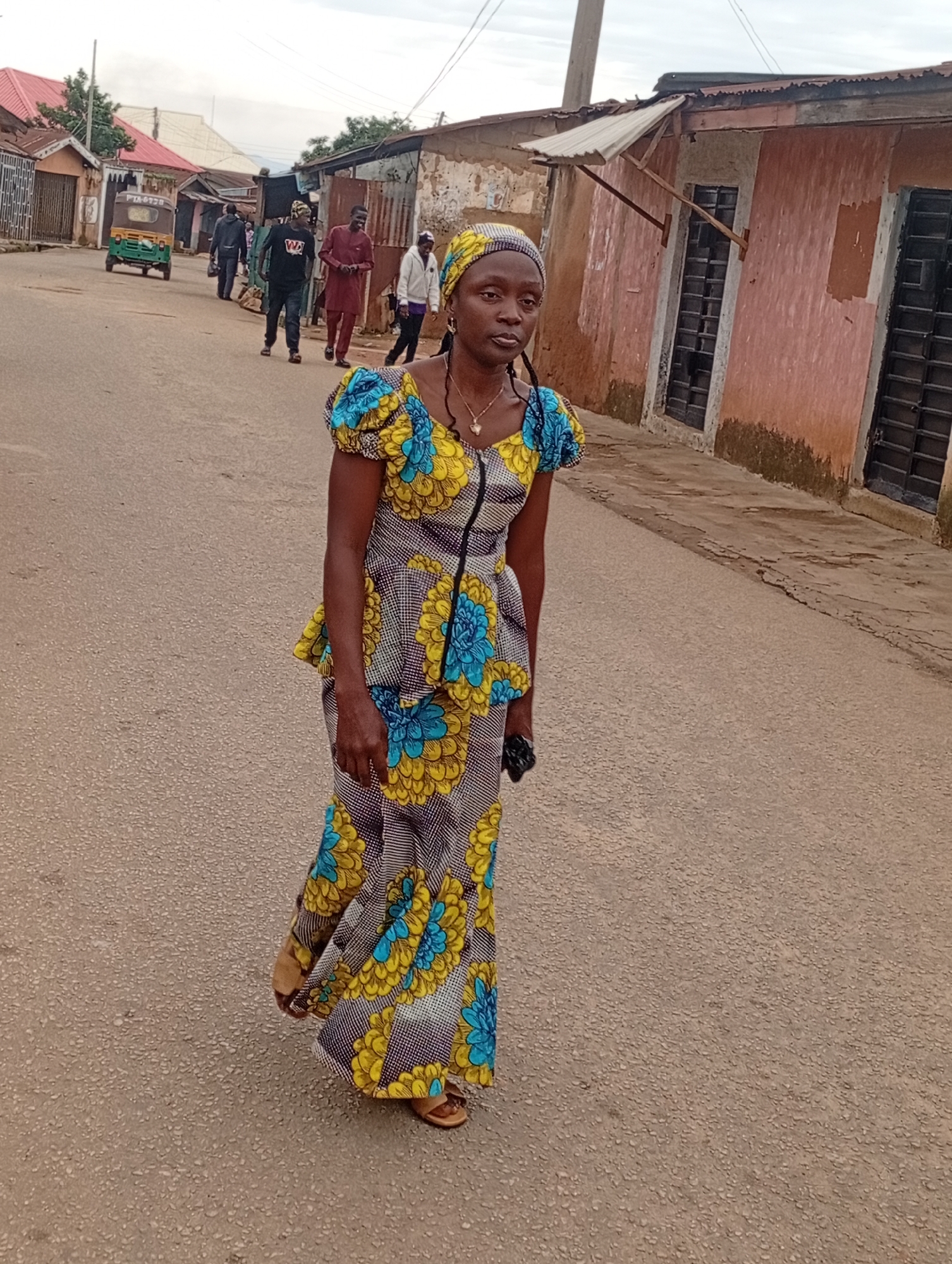 In the city of Unknown, Unknown, a woman with a patterned headscarf and a matching vibrantly colored dress walks on a paved street. The dress features a yellow and blue floral design on a white background with thin black stripes. She wears open-toed sandals. The woman is looking directly at the camera. Behind her, several other people are visible walking in the street. To the left of the street, there are buildings with corrugated metal roofs and what appears to be a small shop. A green auto-rickshaw is parked further down the street on the left. The buildings on the right side of the street have pinkish-orange facades and dark barred windows. The sky is overcast.