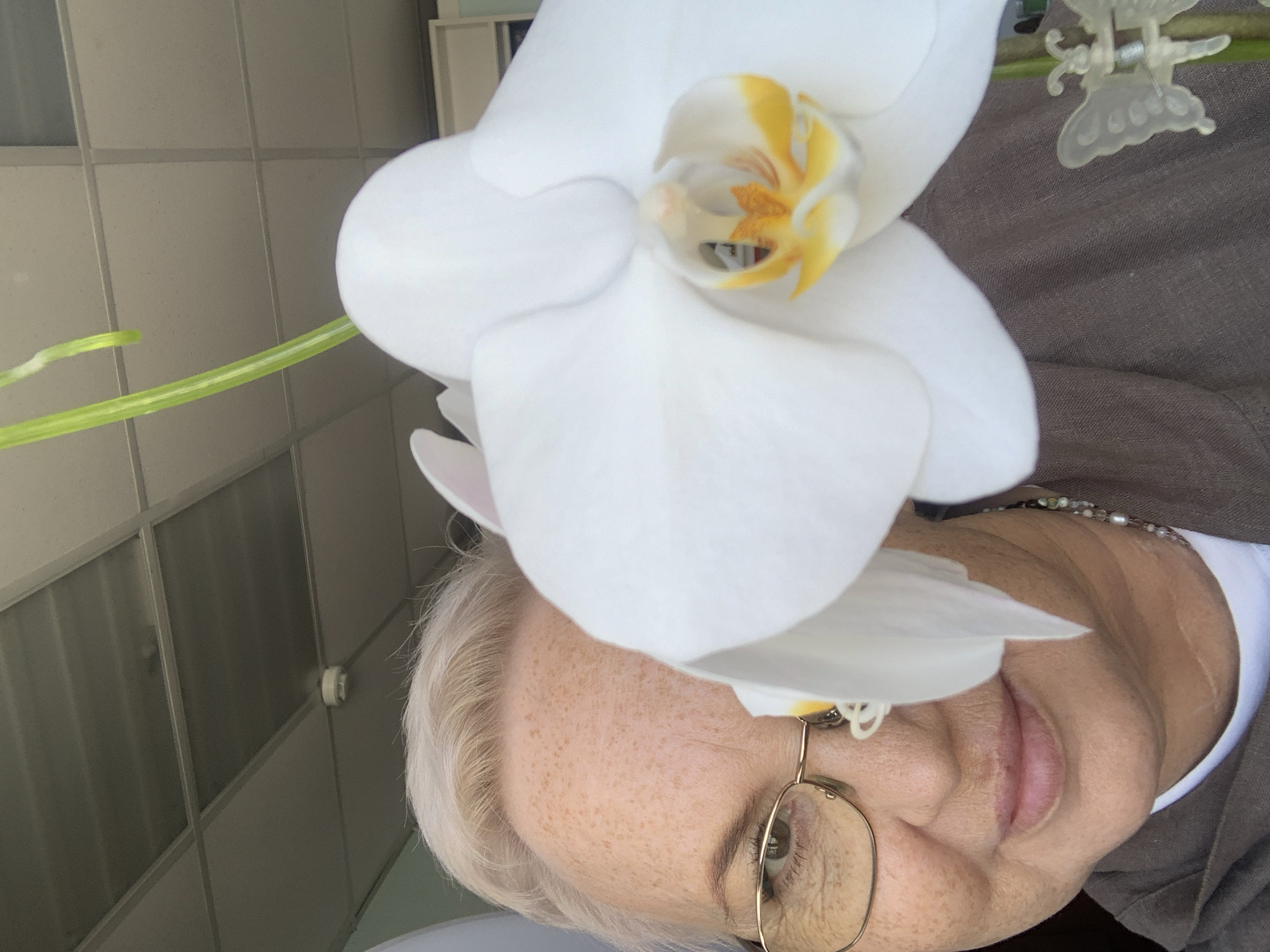 A close-up shot shows a woman with fair skin and short grey hair. She wears glasses with thin gold frames, and a necklace is visible around her neck. A large white orchid flower dominates the frame, partially obscuring her face. The background features a tiled wall and ceiling, suggesting an indoor location. A green stem and a clear plastic butterfly clip are also visible. While the location is specified as Chaikovsky, Russia, no information within the image confirms this city.