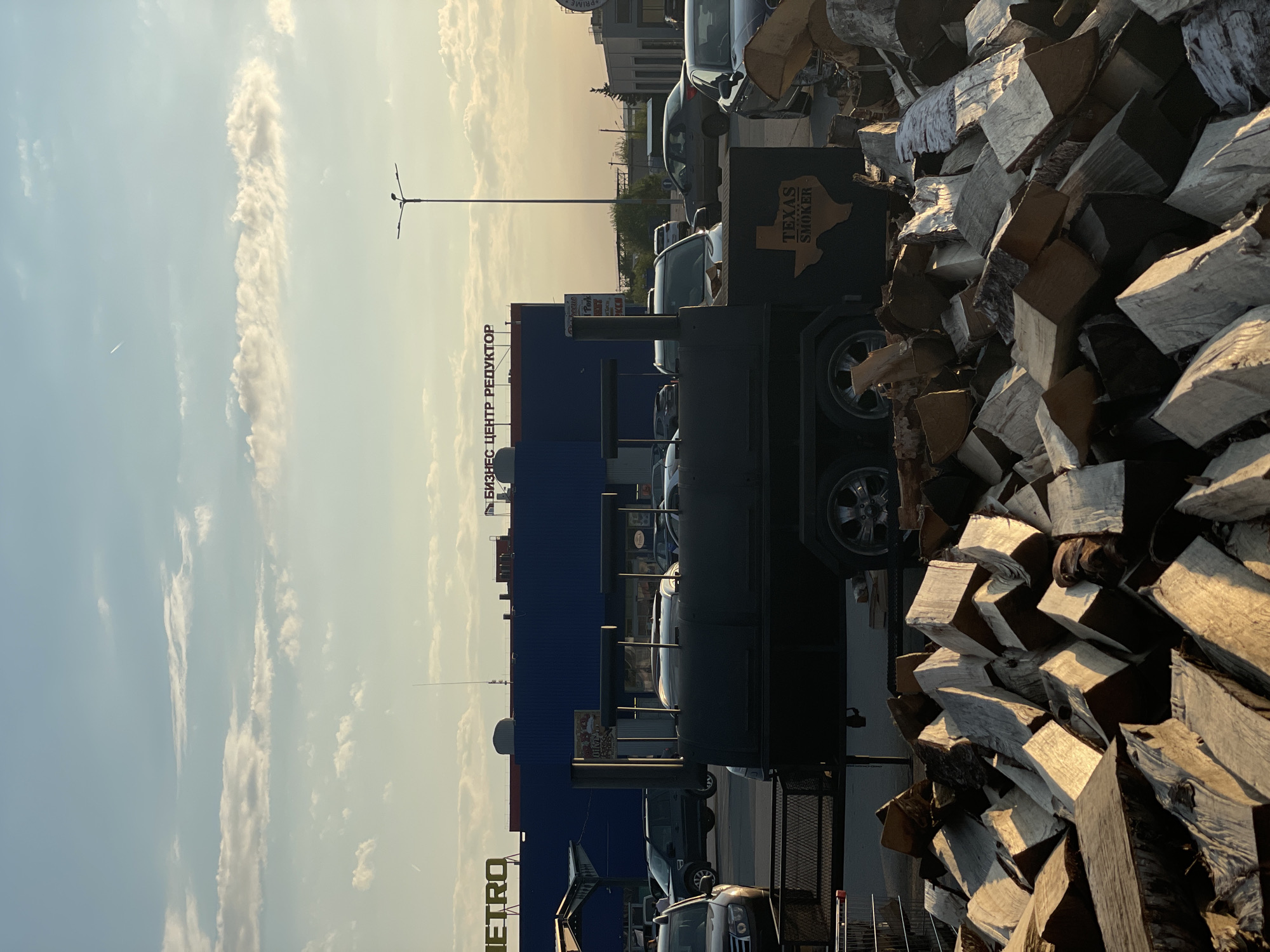 A partially obscured view reveals a "TEXAS SMOKER" smoker on wheels near a pile of logs. Several cars are parked in front of a blue building with "МЕТРО" and "БИЗНЕС ЦЕНТР РЕДУКТОР" signs. The sky is visible above the buildings and is pale blue with white clouds. The scene is outdoors. Based on the Cyrillic text on the building, the location is likely Saint Petersburg, Russia.