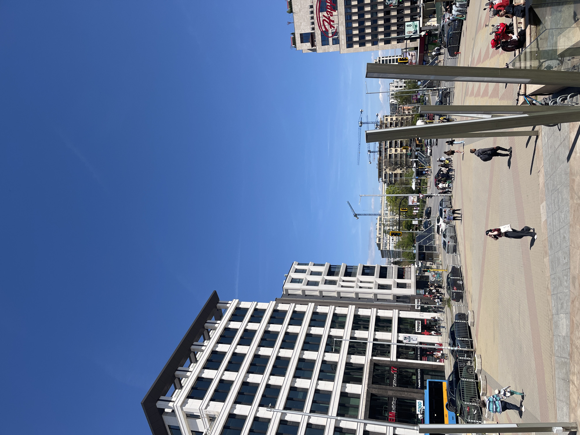The image captures a cityscape in Sofia, Bulgaria, under a clear blue sky. Dominating the view are modern office buildings featuring grid-like facades of white and dark windows.
In the mid-ground, there is a street scene with pedestrian traffic and vehicles. One can see construction cranes in the distance, indicating ongoing development in the area.
In the foreground is a pedestrian walkway paved with light-colored bricks. Several people are walking, and there are parked cars and some motorcycles. There's a bicycle rack and other street furniture visible. Tall, light-colored metal posts or beams are also present.