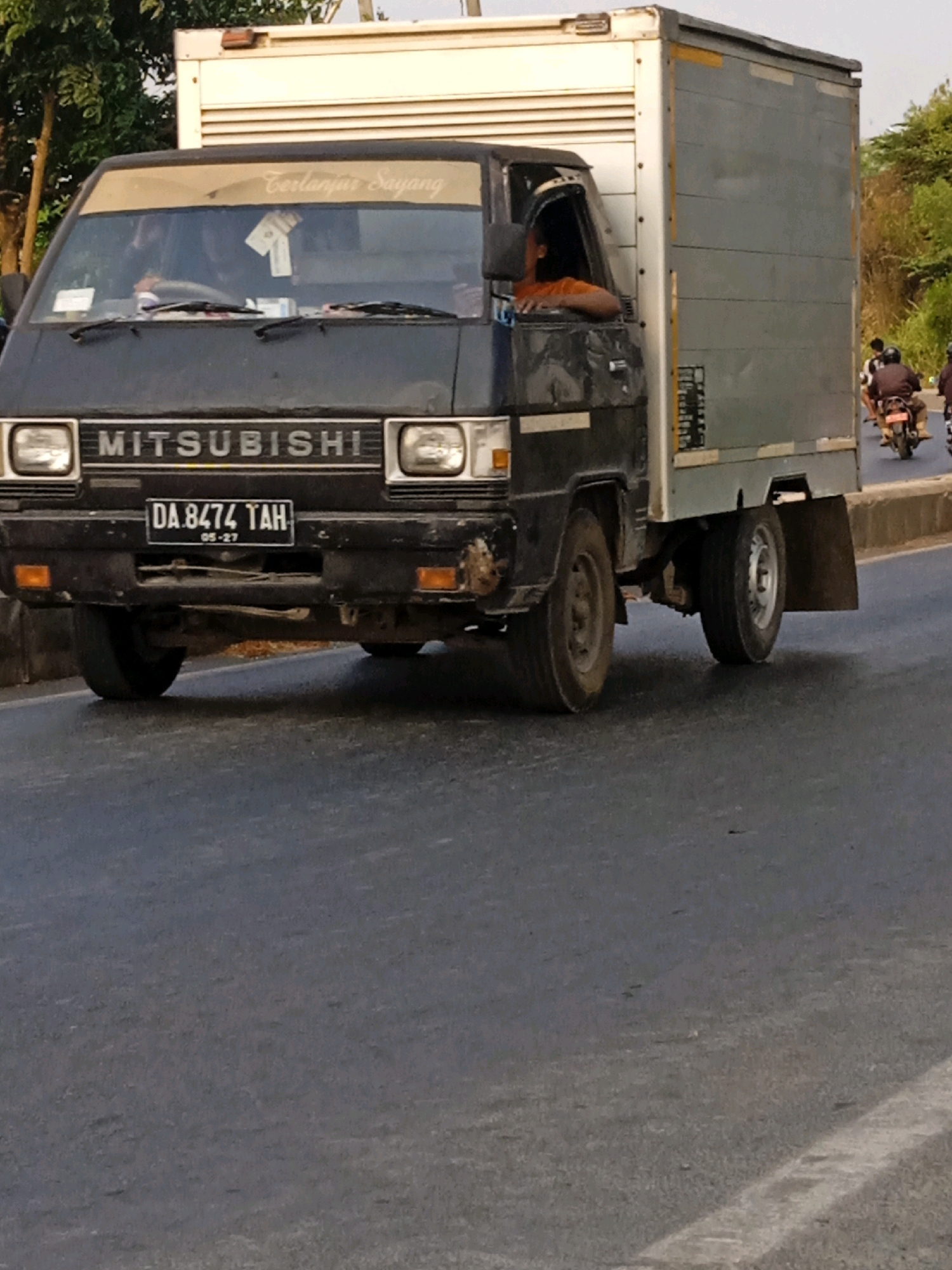 The image shows a dark gray Mitsubishi light truck with a white cargo box, driving on a paved road in Banjarmasin, Indonesia. The truck has a license plate that reads "DA 8474 TAH" with "05-27" below it. A person, wearing an orange shirt, is visible in the driver's seat, looking out the window. Another person is partially visible in the passenger seat. The truck's windshield has a sticker that says "Terlanjur Sayang". In the background, there are trees and other vehicles, including motorcycles. The road appears to be a city street with some foliage and buildings visible on the side. The lighting suggests it is daytime.