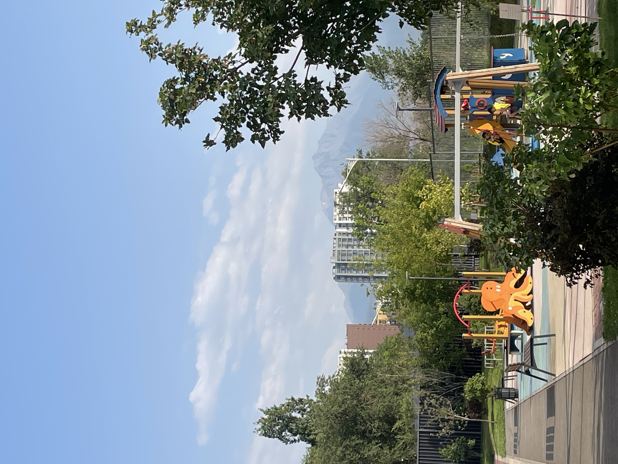 This image depicts a modern playground situated in Almaty, Kazakhstan, with a backdrop of the city's skyline and the majestic Tien Shan mountains. The playground features a variety of colorful equipment, including a large orange octopus-shaped slide, swings, and climbing structures. Two children are visible on the playground, one on a slide and another sitting on a structure. Lush green trees and bushes surround the play area, suggesting a well-maintained urban park or residential complex. In the distance, several multi-story buildings are visible, indicative of urban development, and the imposing silhouette of the Tien Shan mountains provides a stunning natural contrast to the city's architecture. The sky is a clear blue with scattered white clouds, suggesting fair weather.