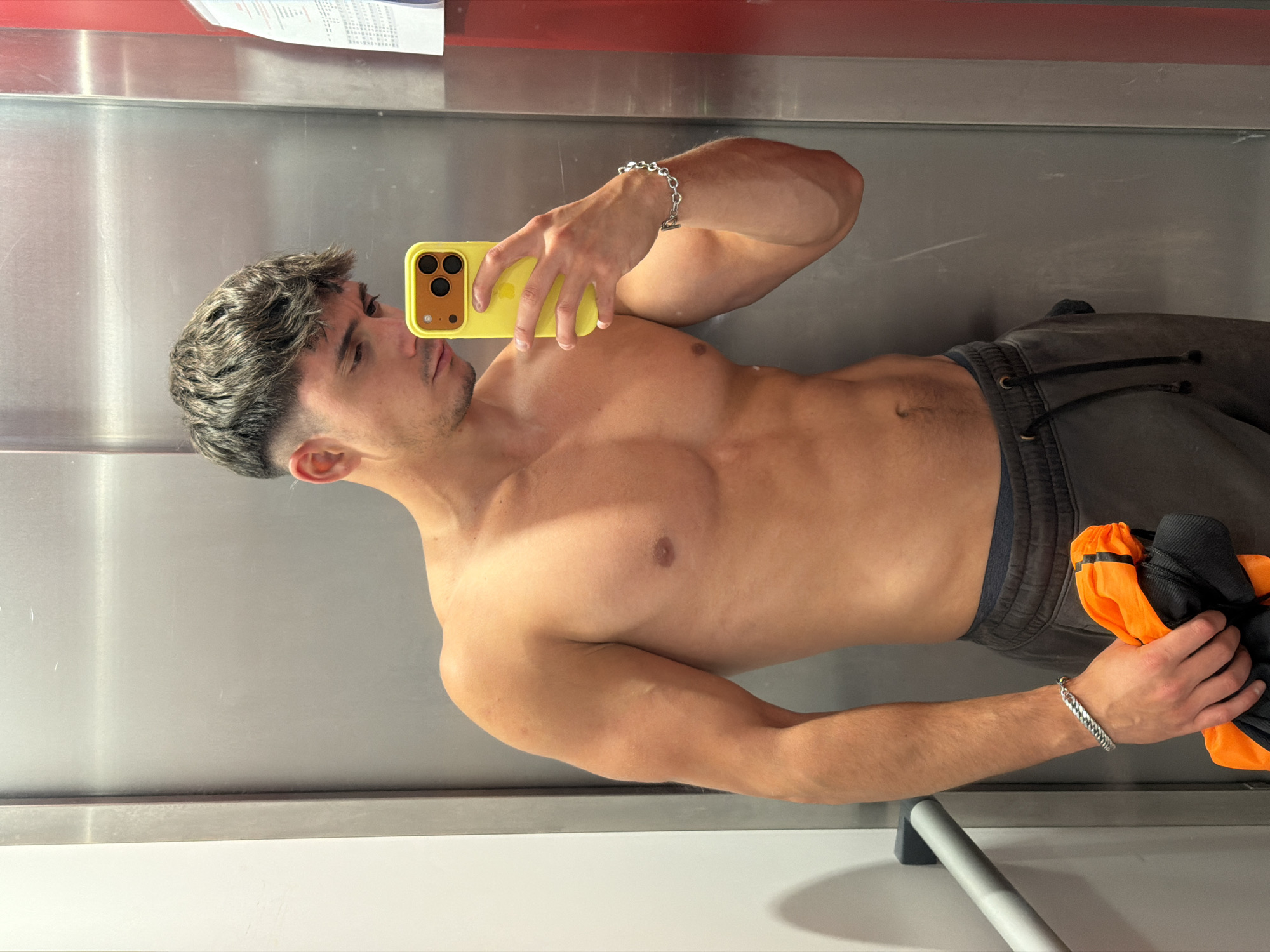 A shirtless young man with a fade haircut and a slight stubble takes a mirror selfie in an elevator. He is wearing dark gray sweatpants and has a silver chain bracelet on his left wrist. In his right hand, he holds a bright orange garment. The elevator walls are metallic gray, and a white wall with a gray handrail is visible to the left. A red vertical stripe is present on the right side of the image, and a piece of paper is affixed to it. The lighting appears to be artificial and casts shadows on the man's torso. The scene is consistent with an elevator in the city of Sofia, Bulgaria, as indicated by the specific elevator design and context.