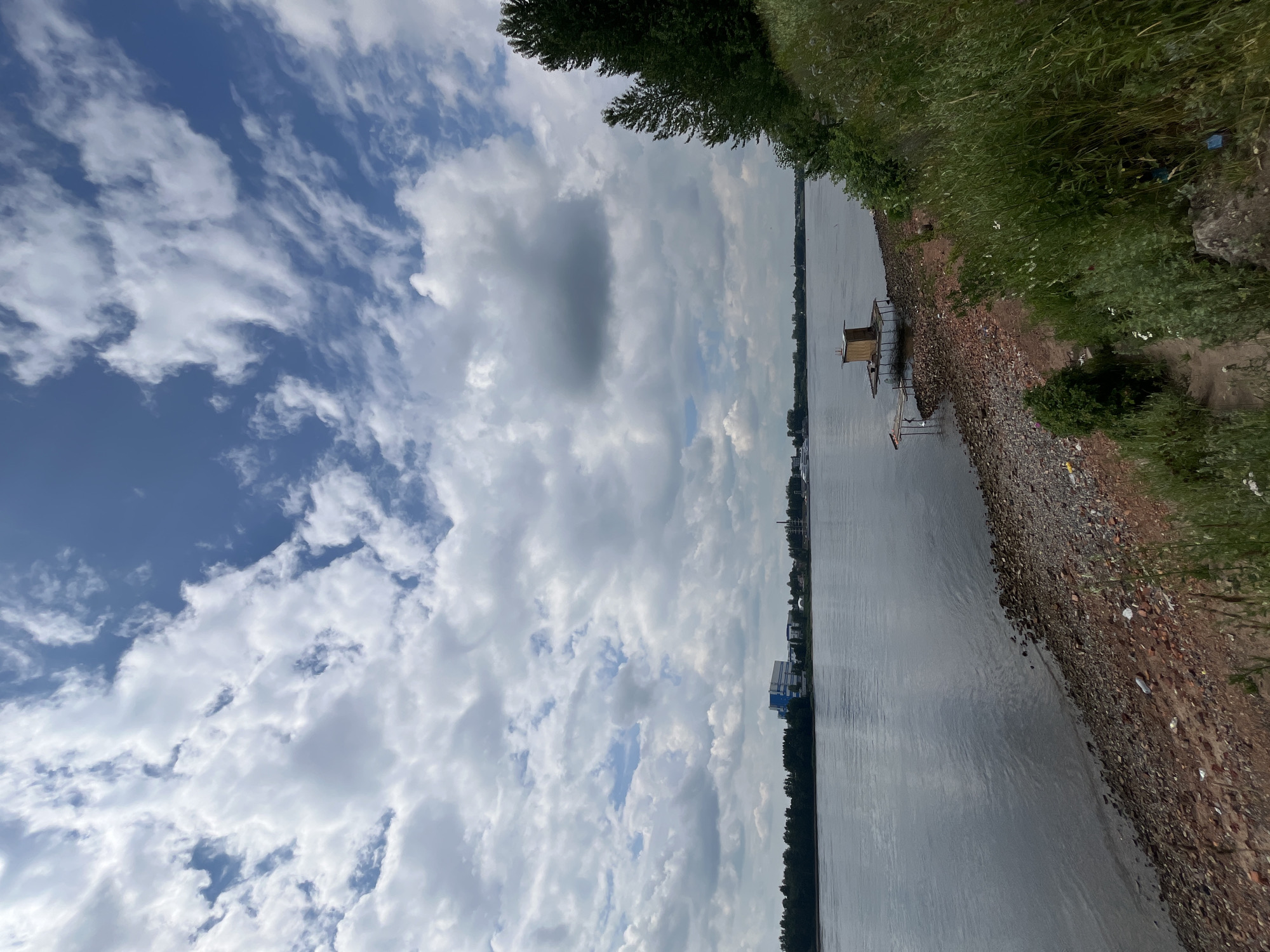 The image shows a wide, calm river under a sky filled with scattered clouds. On the far bank, a line of trees is visible, with some buildings and industrial structures interspersed. In the foreground, the riverbank is composed of stones, sand, and some scattered debris, including pieces of brick and plastic. A small wooden structure, possibly a fishing hut, is situated on a platform extending into the water, connected by a wooden walkway. Lush green vegetation grows on the right side of the bank, suggesting a natural, undeveloped area. The scene appears to be taken on a sunny, possibly overcast day. The location is identified as being in the city of Imeni Sverdlova, Russia.