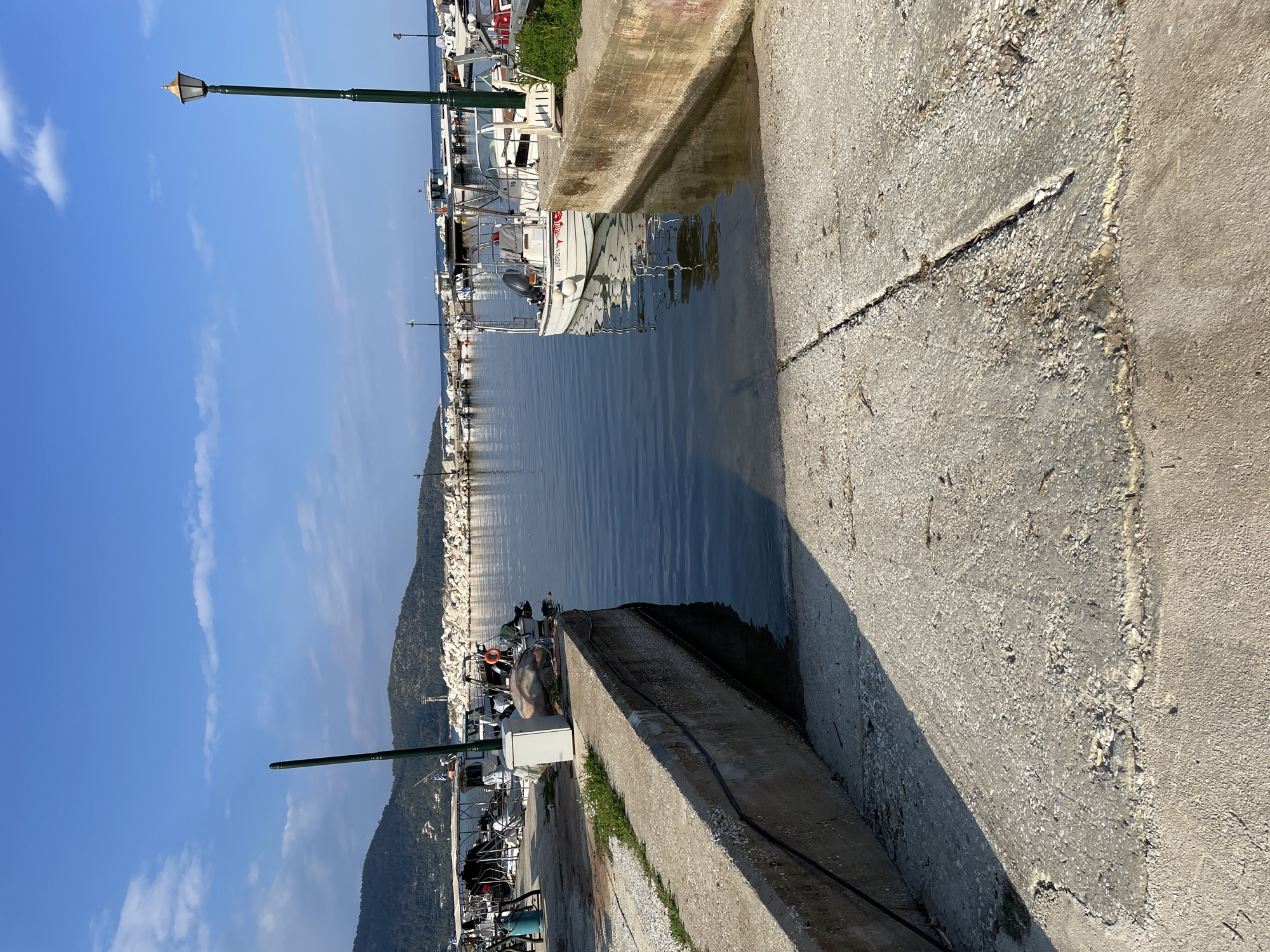 This image depicts a waterfront view in Skala Potamias, Greece, characterized by a concrete pier or dock. Fishing boats are moored along the waterfront. The water is relatively calm. A green lamp post stands adjacent to the concrete structure. A wooded hill is visible in the background. The sky is blue with some clouds. The light suggests it is daytime. The composition emphasizes the texture of the concrete in the foreground.