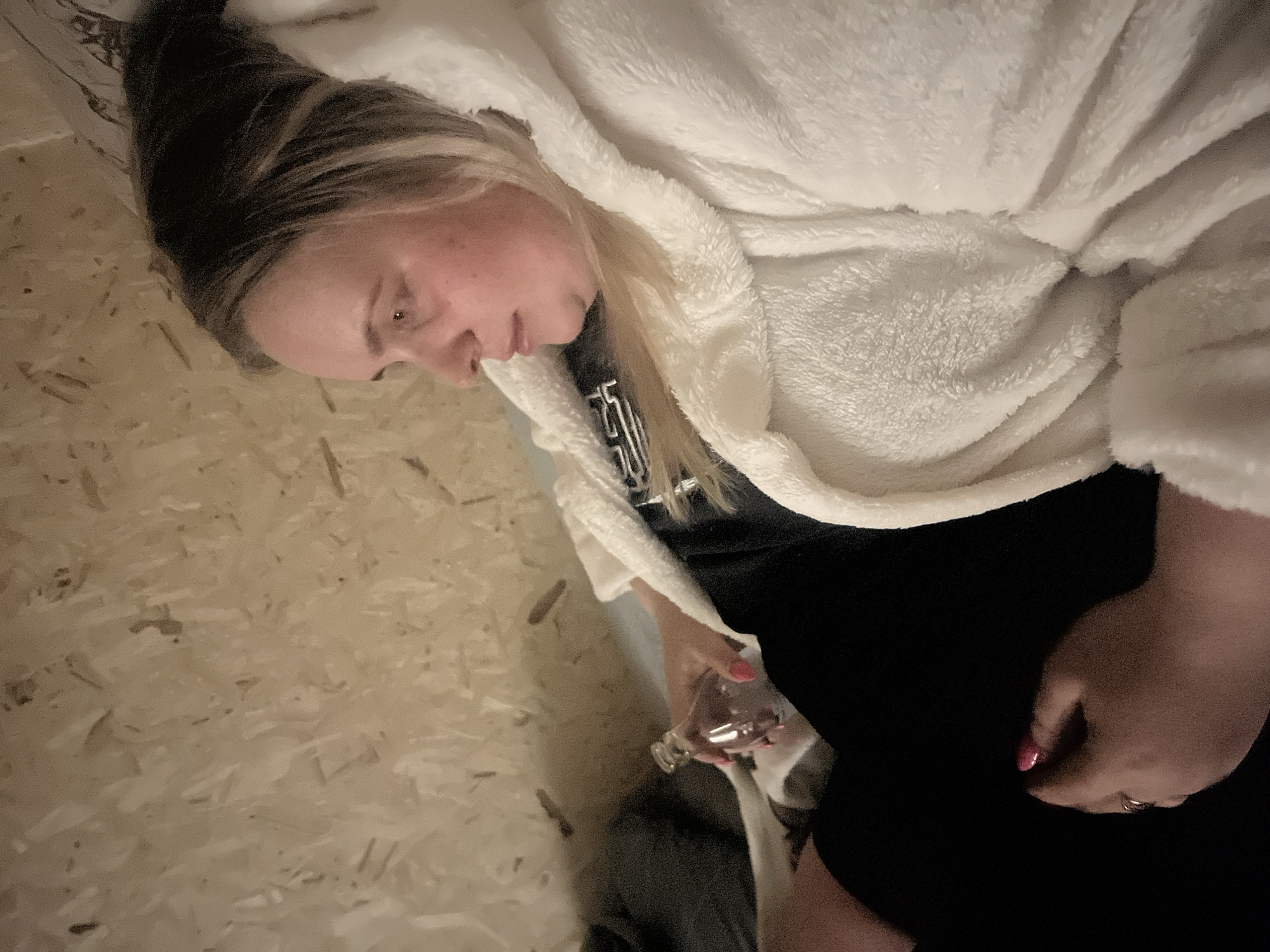 The image shows a woman indoors, possibly in a room with wood paneled walls. She is wearing a white, fluffy robe over a dark shirt and pants. She is holding a small, clear glass in her left hand. The woman has blonde and dark hair and is wearing a ring. The location is indicated as Chaikovsky, Russia, though the photograph itself does not offer any contextual clues. The woman appears relaxed, but her specific activity is not definitively clear.