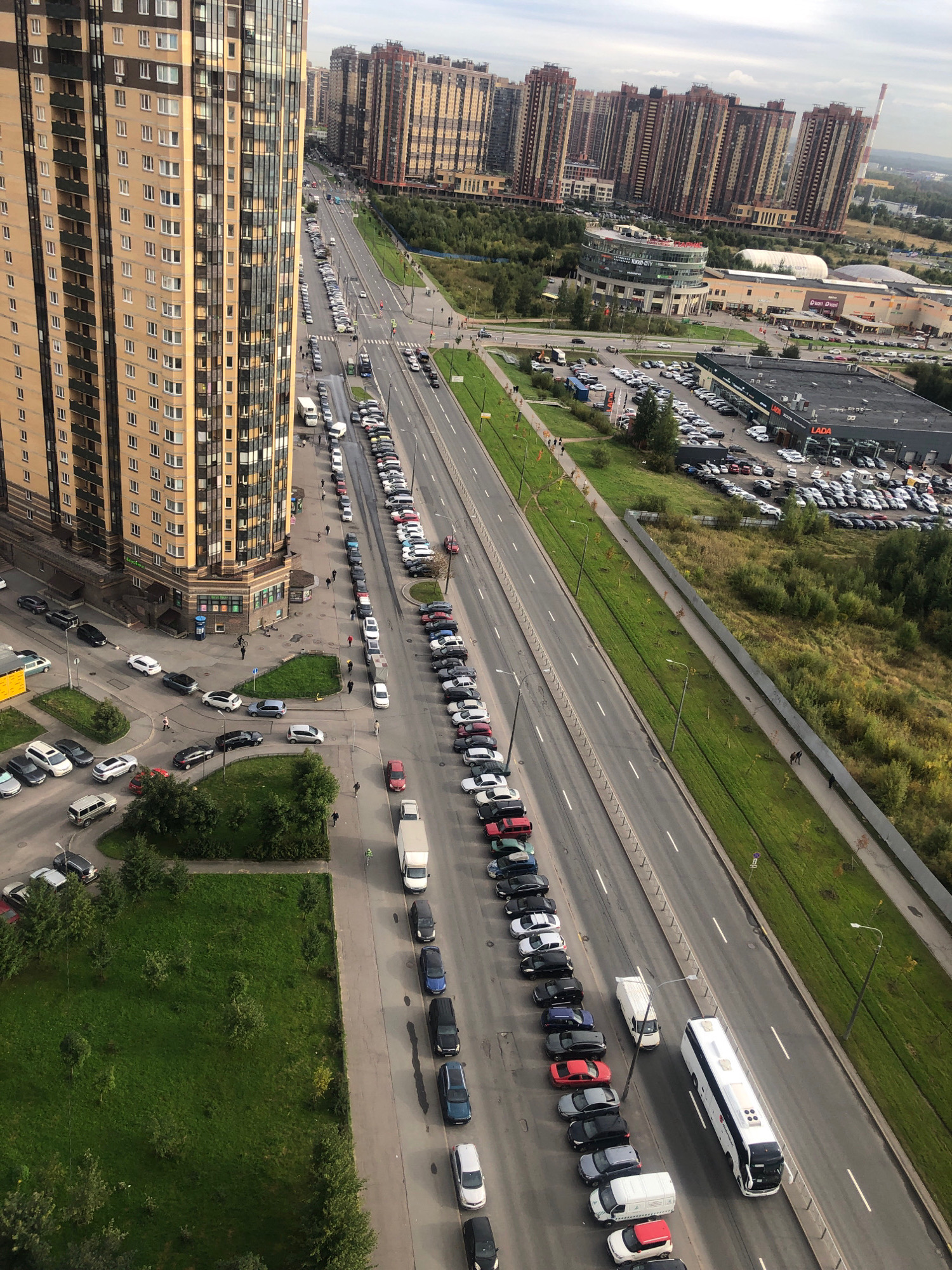 The aerial image captures a wide view of a suburban or outskirt area of an unknown city in Russia. A prominent, tall, multi-story residential building with a beige and dark-paneled facade dominates the left side of the frame. Adjacent to this building is a multi-lane asphalt road, with cars parked in rows along the right side and some in traffic lanes. The road runs parallel to a strip of green lawn and then a dense area of trees and vegetation. Across the road and further into the distance, a collection of similarly styled, tall residential buildings are visible, suggesting a large-scale housing development. Beyond the buildings, a commercial area is discernible with a large building labeled "LADA" which appears to be a car dealership, with many cars parked in its lot. The sky is overcast, suggesting a cloudy day. Several people are seen walking on sidewalks and near the buildings. The scene depicts a typical urban or semi-urban environment with residential and commercial infrastructure.