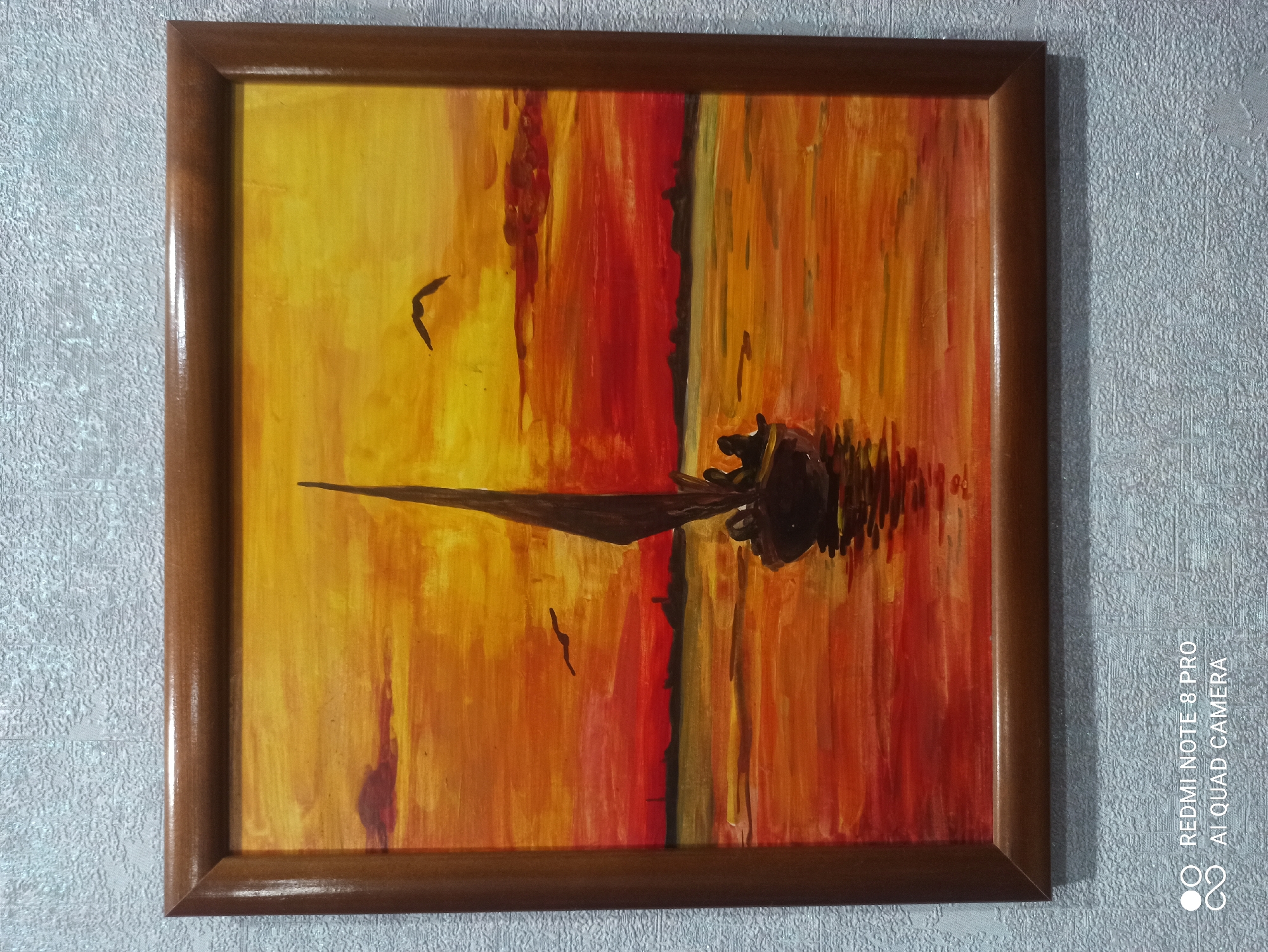 The image displays a framed painting depicting a sunset over a body of water with a sailboat. The sky is a vibrant gradient of yellow, orange, and red, with two birds flying in the distance. A silhouetted shoreline with indistinct structures is visible on the horizon. The water reflects the fiery colors of the sunset. A small sailboat with a dark sail and several silhouetted figures aboard is positioned in the foreground, slightly to the right of center. The painting is framed by a simple wooden frame. The background of the image is a textured grey wall. No information about the city of Kugesi, Russia is present in the image itself.