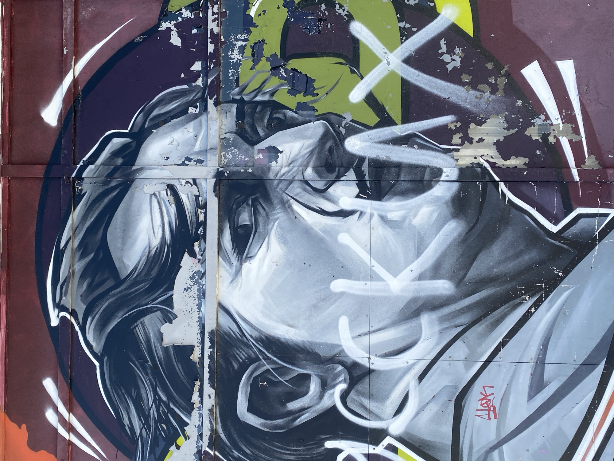 A close-up photograph depicts a section of a graffiti-covered wall in Заволжье, Russia. The focal point is a monochrome portrait of a person with their head tilted back. The artwork is marked with white graffiti lines. The background features layers of peeling paint in shades of maroon, purple, and green. The wall is constructed of metal panels, with vertical seams clearly visible. A signature in red ink is present in the lower right portion of the image. The paint is peeling and cracked throughout the image.