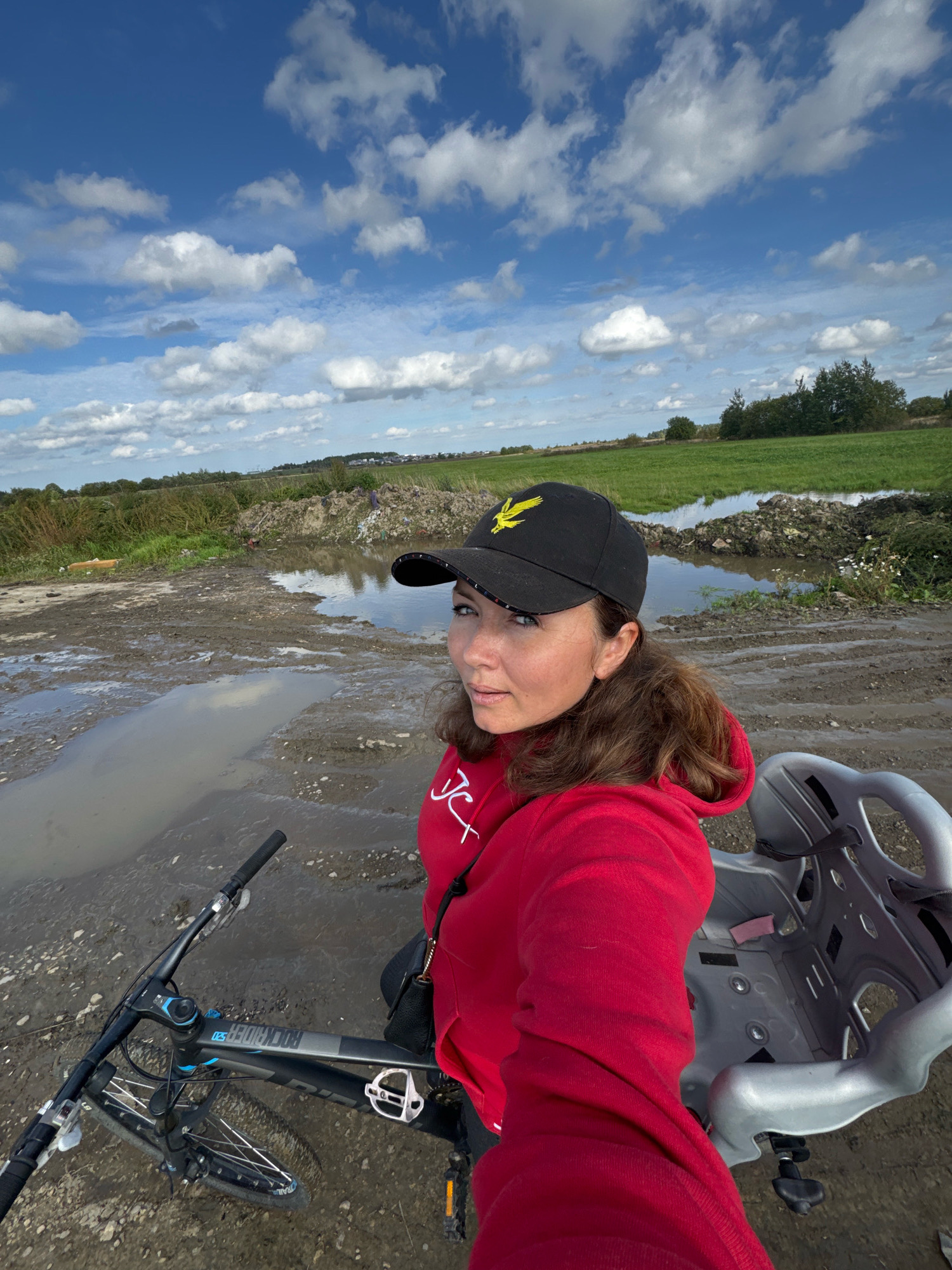 In Kempelevo, Russia, a woman stands next to a bicycle with a child seat attached. She is wearing a red hoodie and a black baseball cap with a yellow eagle emblem. The ground is muddy and there are several large puddles. In the background, there is a field of green grass, a pile of debris, and trees. The sky is blue with white clouds.