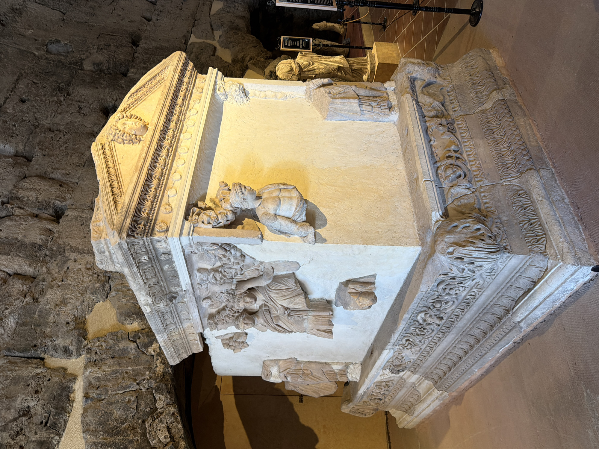 This image depicts a marble sarcophagus, likely of Roman or Byzantine origin, displayed in an indoor exhibition space. The sarcophagus is intricately carved with figures and decorative motifs, including what appear to be human busts and relief sculptures of draped figures. The sarcophagus rests on a richly ornamented base, also of marble. The background reveals a rough, stone-lined wall, suggestive of an ancient structure or cave-like environment, possibly within a museum.
The sarcophagus is located in Hierapolis, an ancient city located on hot springs in southwestern Turkey, near the modern-day city of Denizli. The ruins of Hierapolis are part of the Pamukkale UNESCO World Heritage Site, known for its white travertine terraces formed by thermal waters. The sarcophagus is likely displayed within the archaeological museum of Hierapolis, which houses artifacts excavated from the ancient city and its necropolis. The lighting suggests an artificial indoor environment.