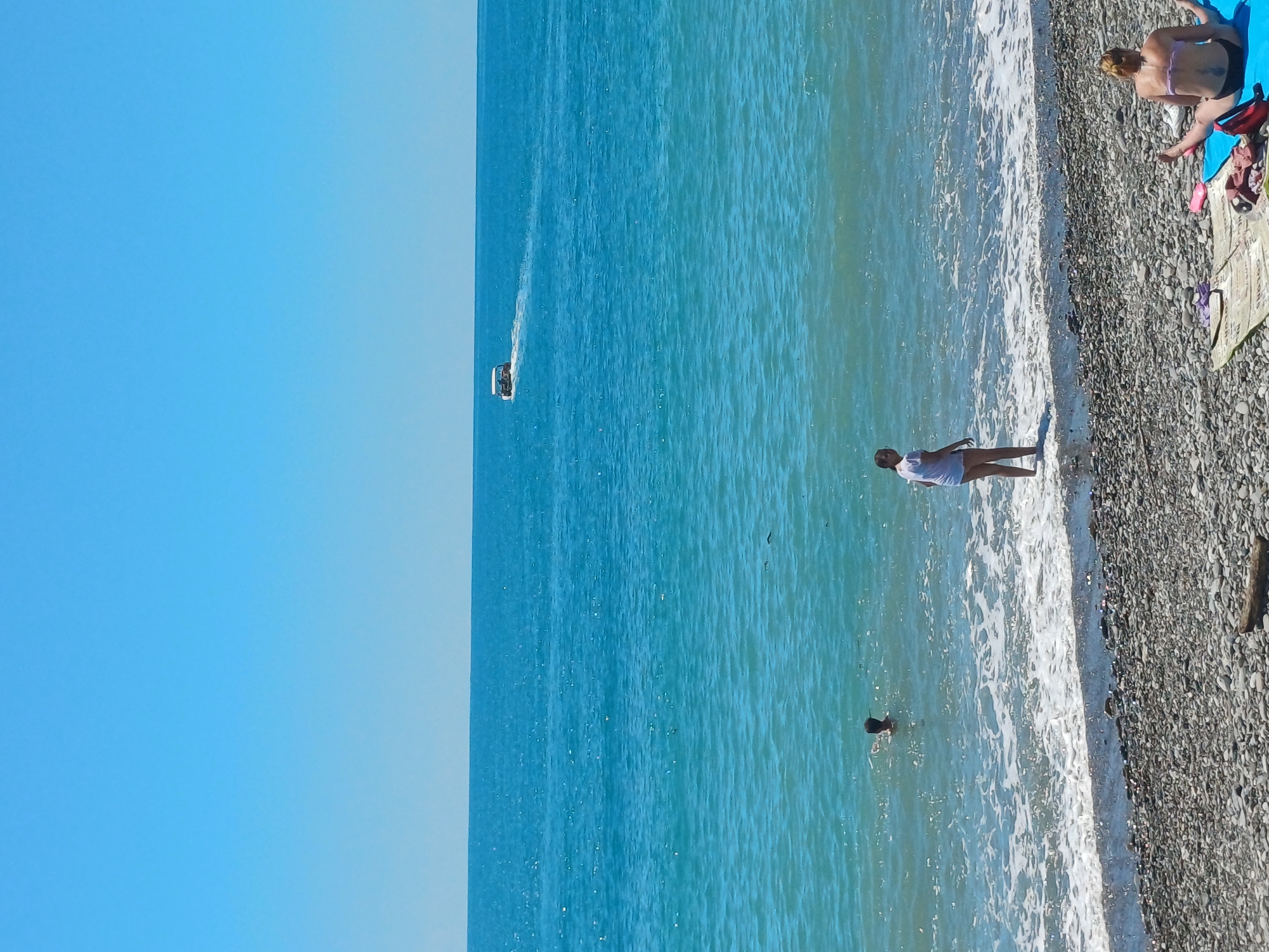 The image depicts a sunny day at a pebble beach in Sochi, Russia. The sea is a vibrant turquoise, stretching out to meet a clear blue sky. A small boat is visible in the distance, leaving a white wake as it moves across the water. In the foreground, several people are enjoying the beach. One person is wading in the shallow water near the shore, while another stands on the pebbles looking out at the sea. Further down the beach, two individuals are relaxing on towels. One person is seated with their legs spread, facing away from the camera, and the other is also seated on a towel. The beach itself is covered in smooth, grey pebbles. The overall atmosphere is one of leisure and summer recreation.