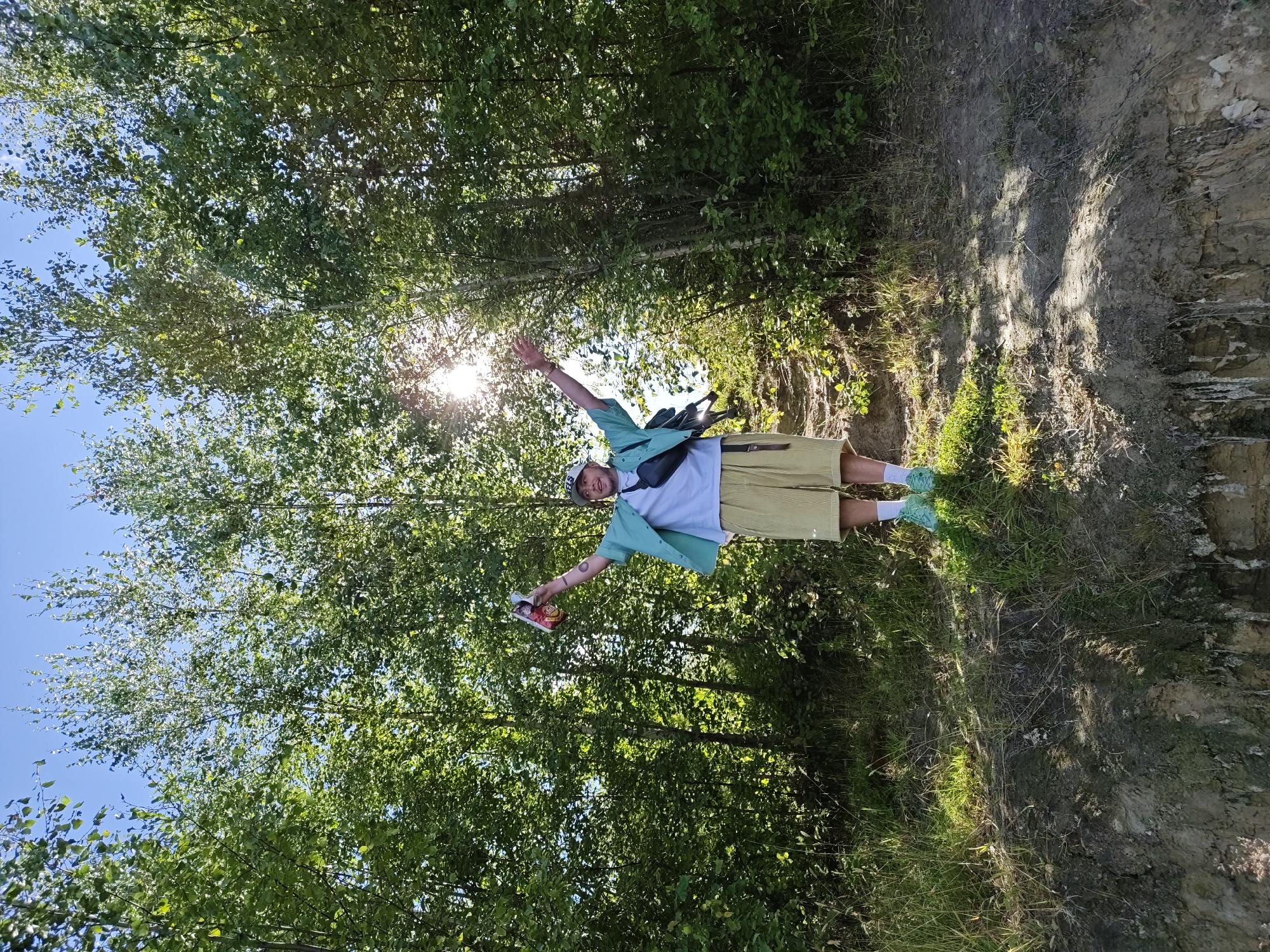 A young man stands in a forest clearing, holding a red can of drink aloft in his right hand. He wears a teal short-sleeved shirt over a white t-shirt, a black fanny pack across his chest, light yellow shorts, white socks, and bright green sneakers. He has a white baseball cap on and is looking up towards the sun, with his arms spread wide. The sun is shining brightly through the trees, creating a lens flare. The ground in front of him is a steep, eroded slope with dirt and some greenery. The background is dense with trees. This scene appears to be located in a natural park or wooded area, potentially in the vicinity of Toksovo, Russia, given the characteristic birch trees visible.