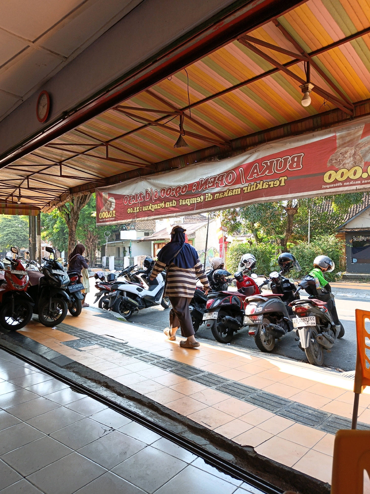 The image depicts an outdoor scene in Klaten, Indonesia, likely near a street-side establishment. A red and white banner with text in Indonesian hangs from the awning of a building, which features a multi-colored striped fabric roof in yellow, orange, green, and pink. Several motorcycles are parked along the sidewalk in front of the building. One woman wearing a headscarf and a striped shirt walks away from the viewer. Another woman in a dark headscarf stands near a red motorcycle. The ground is paved with light brown square tiles and a grey tactile paving strip. Sunlight casts shadows, indicating it is daytime. A red clock is visible on the wall of the building to the left. The overall atmosphere suggests a commercial area.