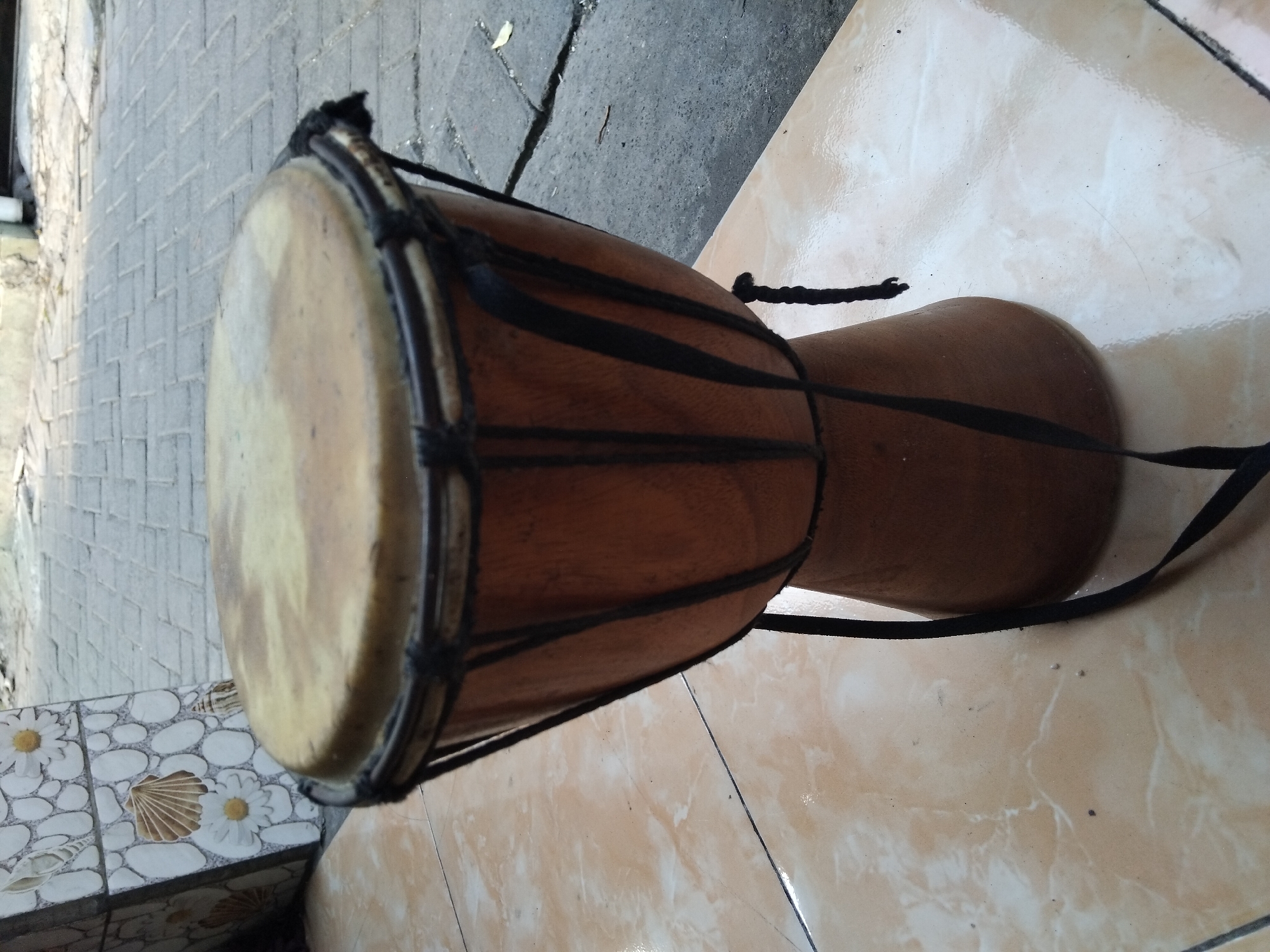 A djembe drum, made of wood with a skin head, rests on a tiled floor outdoors. The drum is brown and has black rope lacing holding the skin head in place. A black strap hangs from the drum. In the background, a wall decorated with mosaic tiles of shells and flowers is visible, as well as a paved walkway. The scene appears to be in an urban environment, possibly a courtyard or an alleyway, located in the city of Kepuh Kiriman, Indonesia. The lighting suggests it is daytime.