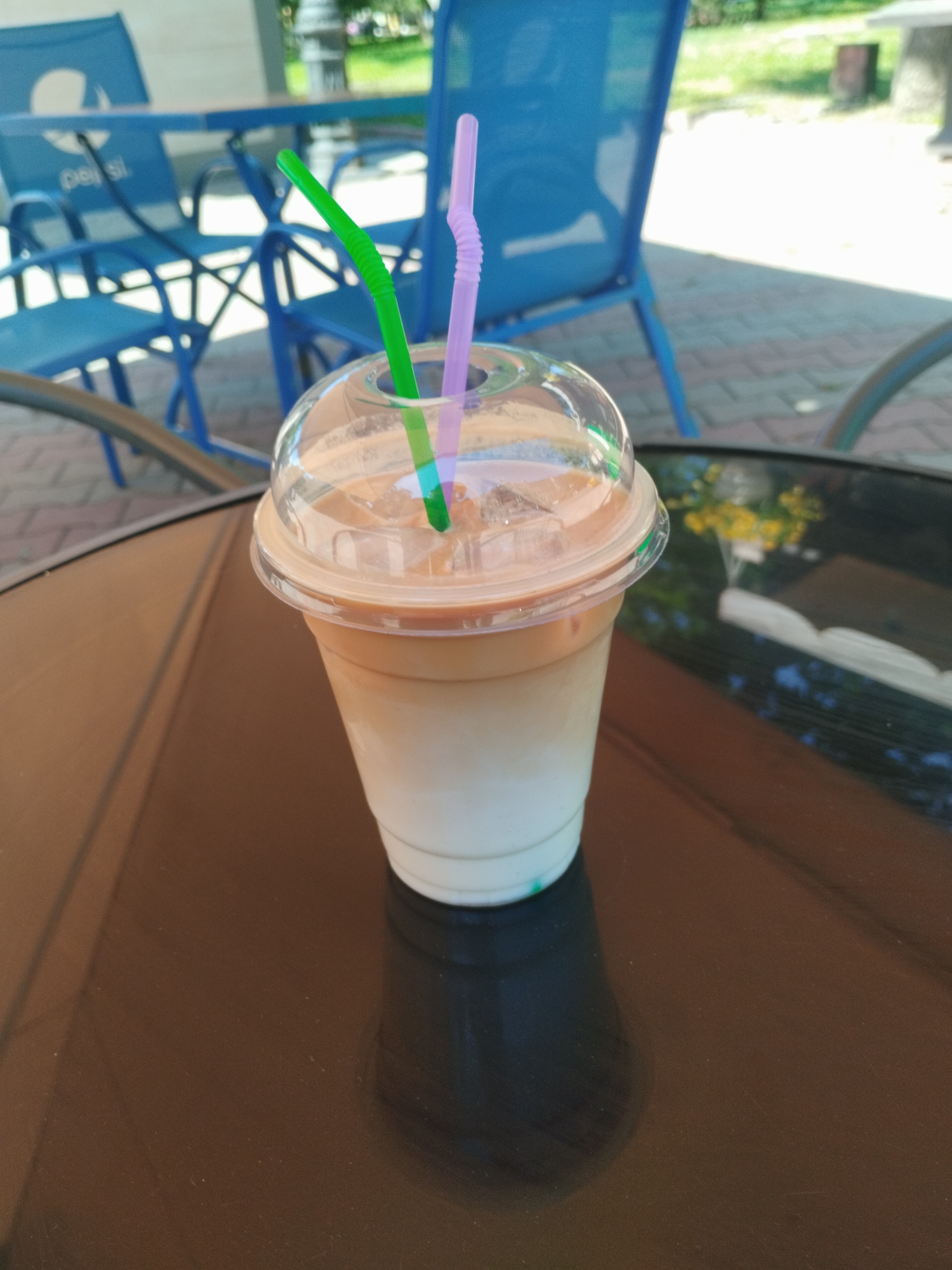 A tall clear plastic glass with a lid filled with a layered beverage, possibly iced coffee, and containing two straws, one green and one purple, sits on a dark reflective tabletop. The drink is lighter in color at the bottom, gradually becoming darker at the top. The tabletop appears to be part of an outdoor cafe setting, as indicated by the presence of blue metal patio chairs with the Pepsi logo visible in the background. The chairs are positioned near a table on what seems to be a patio paved with reddish-brown bricks. In the distance, there's a hint of greenery, suggesting a park or garden. This image was taken in Odesa, Ukraine.