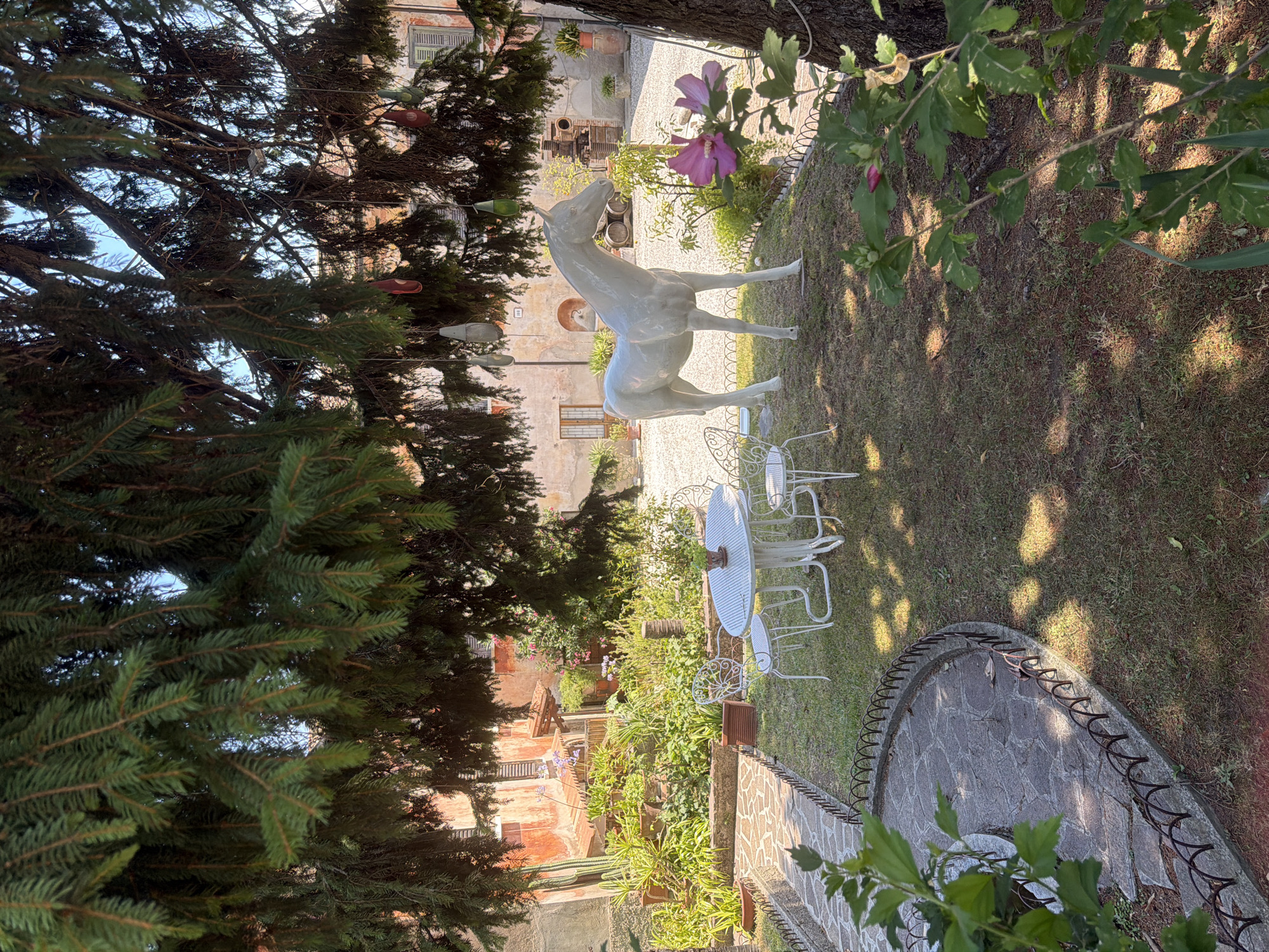 The image depicts a serene garden scene with a white horse statue as the focal point. The statue is positioned on a grassy lawn, surrounded by lush greenery and flowering plants, including a prominent pink hibiscus. In the background, an old building with a terracotta-colored facade and shuttered windows suggests a rustic Italian setting. A white wrought-iron table and chairs are set up on the lawn, inviting relaxation. The garden is framed by the branches of a large evergreen tree, casting dappled sunlight on the ground. A winding stone pathway leads into the garden, bordered by a low metal fence. The overall impression is one of tranquility and rustic charm. The context provided indicates that this scene is located in Padenghe sul Garda, Italy.