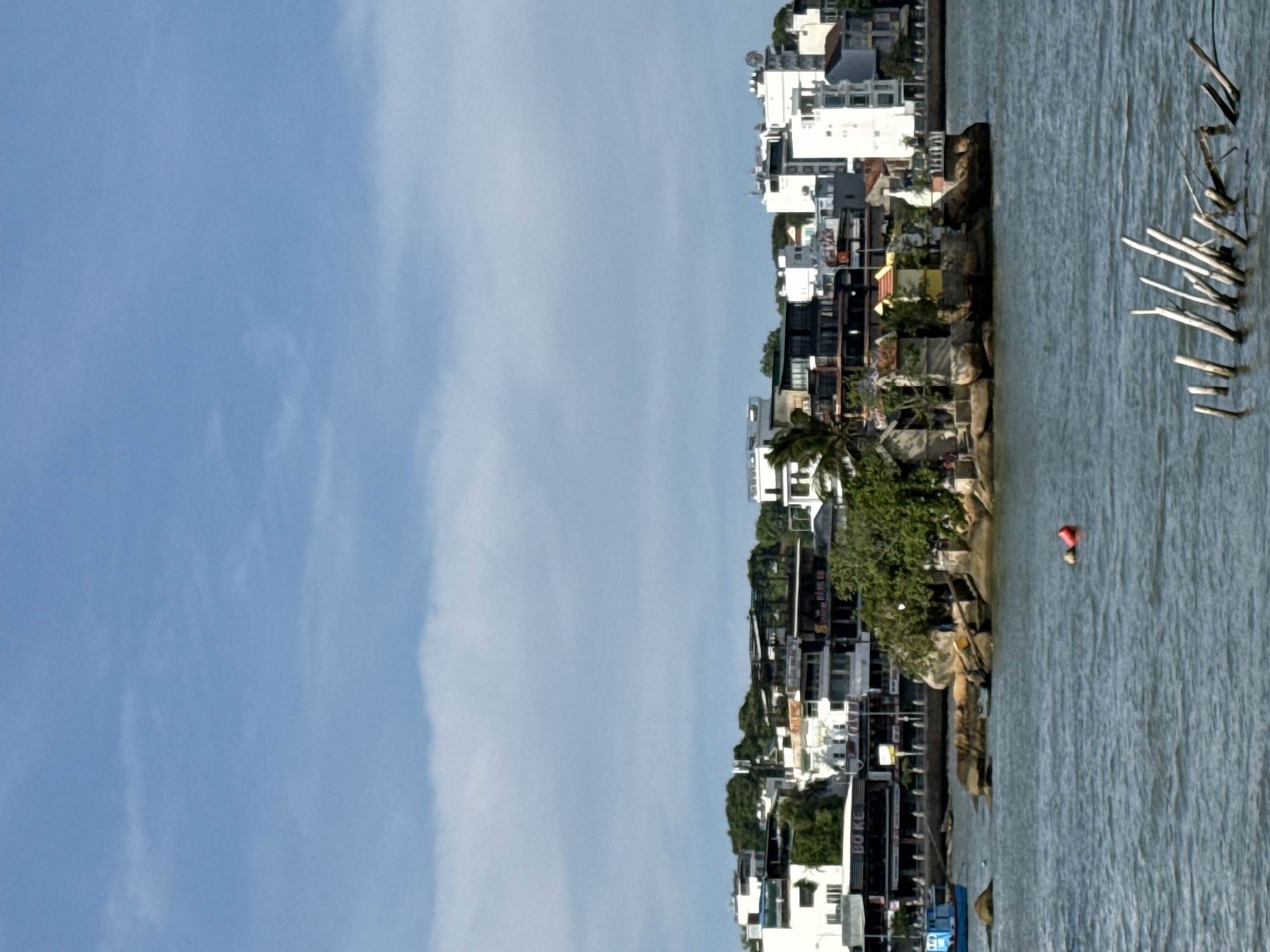 In the city of Bac Nha Trang Ward, Vietnam, a body of water with moderate waves reflects the light from a partly cloudy sky. In the foreground, several wooden posts emerge from the water, some partially submerged. Further out, a small island is visible, densely covered with trees and featuring small structures. There are two red buoys floating on the water near the island. To the left of the island, a blue boat is docked. In the background, a coastal city skyline is apparent, characterized by various buildings of different heights and architectural styles, with a backdrop of lush green hills.