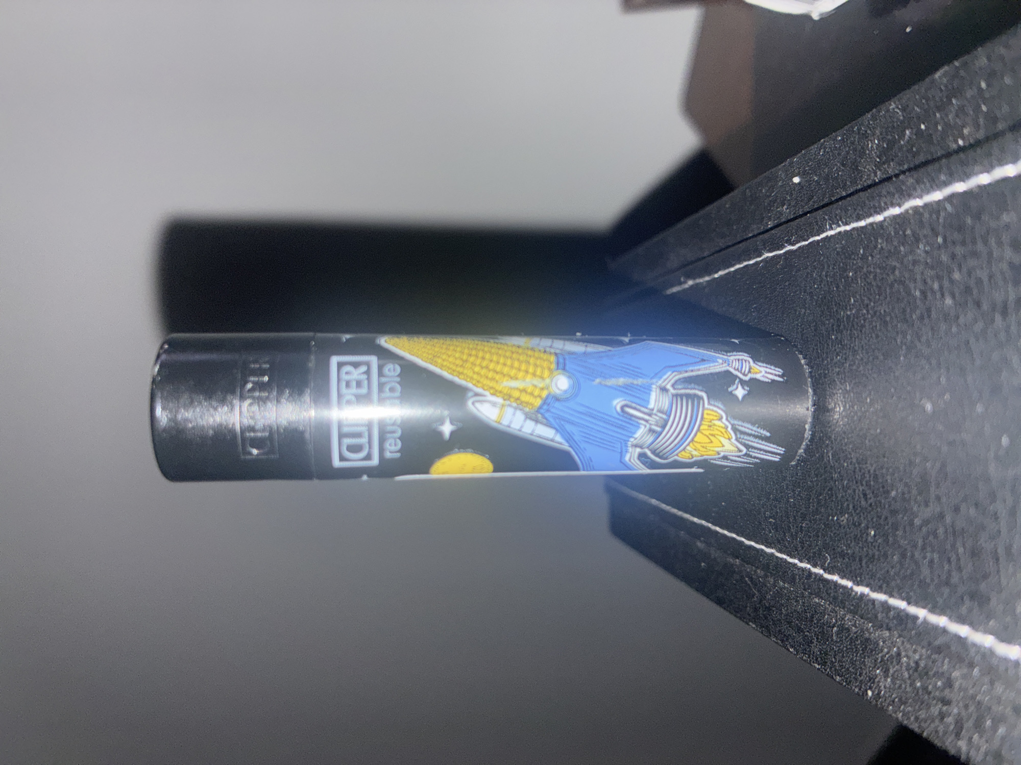 The image shows a black Clipper lighter with a spaceship design lying on a black surface. The lighter is cylindrical and displays the word "CLIPPER" and "reusable." The spaceship is predominantly blue and yellow. The background is out of focus, but appears to be a room with light-colored walls. There is no information available in the image to suggest the location is in Huixquilucan de Degollado, Mexico.