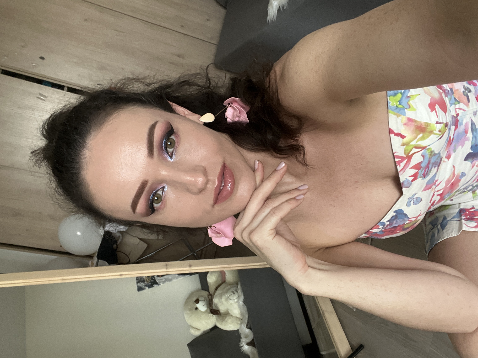 A woman with dark hair and dramatic eye makeup, featuring pink and blue eyeshadow and winged eyeliner, is posing for a selfie. She is wearing a strapless floral dress and large pink petal-shaped earrings. Her right hand is gently touching her chin. In the background, a full-length mirror reflects a beige teddy bear sitting on a grey sofa. The room appears to be a bedroom or living space with wooden paneling on the wall. The image does not provide any specific details to indicate its location in Kazan, Russia.