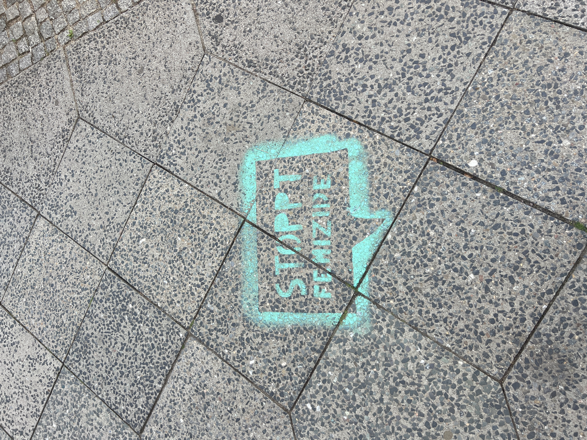 The image shows a section of grey concrete paving stones with a turquoise stencil spray-painted onto them. The stencil depicts a speech bubble containing the words "STOP FEMIZID". The paving stones are part of a sidewalk, with a small section of cobblestones visible at the top right corner. The image is taken from a high angle, looking down onto the ground. There are no people or other distinguishing features visible in the image that would allow for a precise location within Berlin, Germany.