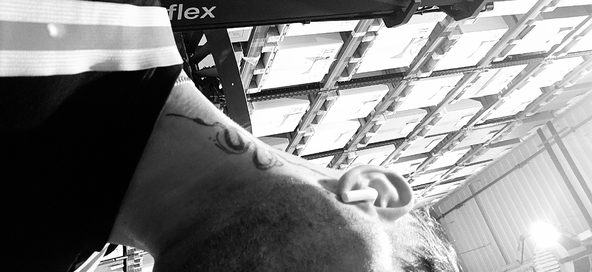 A black and white, low-angle photograph captures a close-up of a person's head and upper body, looking upwards. The subject, positioned on the left side of the frame, has short facial hair and wears an earplug in their visible right ear. A tattoo featuring abstract, curvilinear designs is prominent on their upper arm or shoulder. The person is dressed in dark clothing with a distinctive light-colored, striped band near the top.
The background reveals an industrial or warehouse interior, characterized by an extensive system of metal shelving units. These shelves are densely packed with numerous light-colored boxes or containers, extending into the distance overhead. A dark structural beam, visible in the upper background, features the partial text "flex". The scene is illuminated by artificial lighting, creating a high-contrast aesthetic with bright highlights and deep shadows. The earplug suggests a noisy environment, consistent with an industrial setting. The image's location context is Plovdiv, Bulgaria.