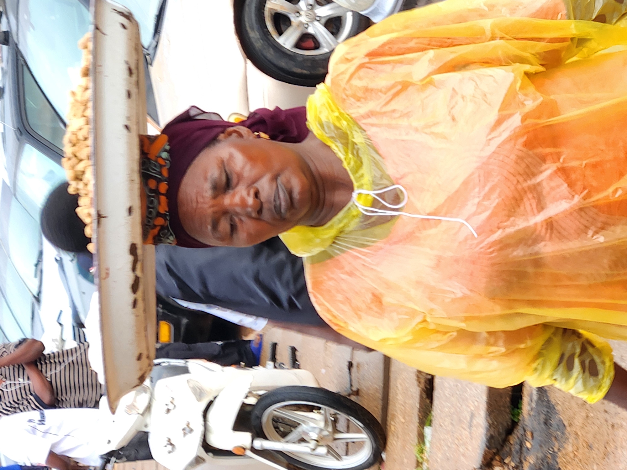 A woman in a yellow poncho and a maroon headscarf balances a tray of food on her head. The tray contains many small, light brown, circular food items. She is wearing a gold hoop earring in her visible ear. She is looking down and to her right, with her eyes closed. In the background, a white scooter is visible to the left. To the right and behind her, the rear wheel and fender of a light-colored car are visible. The ground appears to be paved.