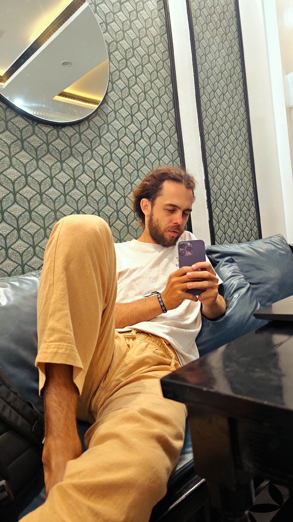 A young man is seated comfortably on a blue sofa in an indoor setting, likely a cafe or lounge in Phố Ràng, Vietnam. He has light brown, curly hair and a beard, wearing a white t-shirt and light brown (khaki) pants. He is engrossed in a purple smartphone, holding it with both hands and looking intently at its screen. A patterned black and white bracelet is visible on his left wrist.
The background features a wall covered in green and white geometric wallpaper, displaying a repeating leaf-like or abstract pattern. An oval-shaped mirror with a black frame hangs on the wall, reflecting a ceiling with warm, recessed lighting and some golden-colored trim. Two dark, vertical structural elements or frames are visible on the wall to the right. A portion of a dark-colored table is visible in the foreground on the right, and a black backpack rests on the sofa near the man's left leg. The man appears calm and focused on his activity. The lighting suggests an indoor setting, and no specific time of day or outdoor weather can be determined. No explicit text is clearly visible, though a faint "N" might be present on the phone's back.