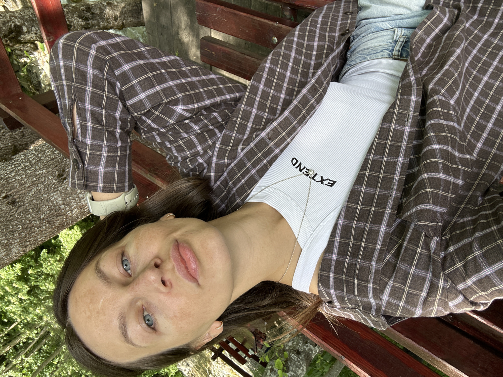 A woman with fair skin, blue eyes, and brown hair is captured from the chest up, sitting on a wooden bench outdoors. She is wearing a brown and purple plaid shirt over a white ribbed tank top with the word "EXTEND" printed in black. She has a delicate silver necklace and small hoop earrings. Her left arm is bent behind her head, and her right arm is resting on the bench. The background consists of a large tree trunk and lush green foliage, suggesting a natural or park-like setting. The lighting indicates it is daytime, with sunlight dappling through the leaves. The image appears to be a selfie, taken at a slightly low angle, with the woman looking directly at the camera. The location is identified as Bare Kraljske, Montenegro.