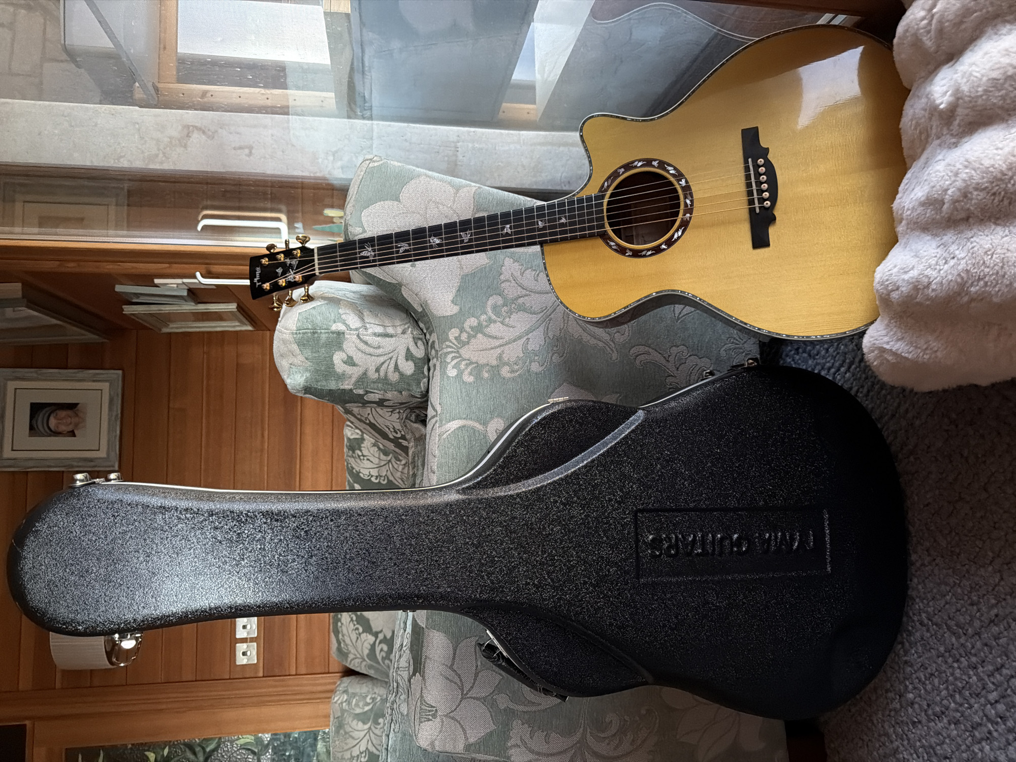 An acoustic guitar case and a blonde acoustic guitar are placed next to each other on a grey carpeted floor in a room with wood-paneled walls and a patterned sofa. The guitar case is black, textured, and stands upright. The guitar is leaning against the sofa, and its neck, headstock, and body are visible. The headstock has gold tuning pegs and a logo that appears to be "TAMA." The fretboard has decorative inlays. The soundhole is surrounded by a decorative rosette. A light-colored, fluffy blanket is in front of the guitar. In the background, framed pictures hang on the wall, and a lamp is visible. A window reflects the outdoors. The scene is indoors in a room that appears to be a living space, possibly in a home in Souni-Zanatzia, Cyprus, though the image alone does not provide definitive proof of this specific location.