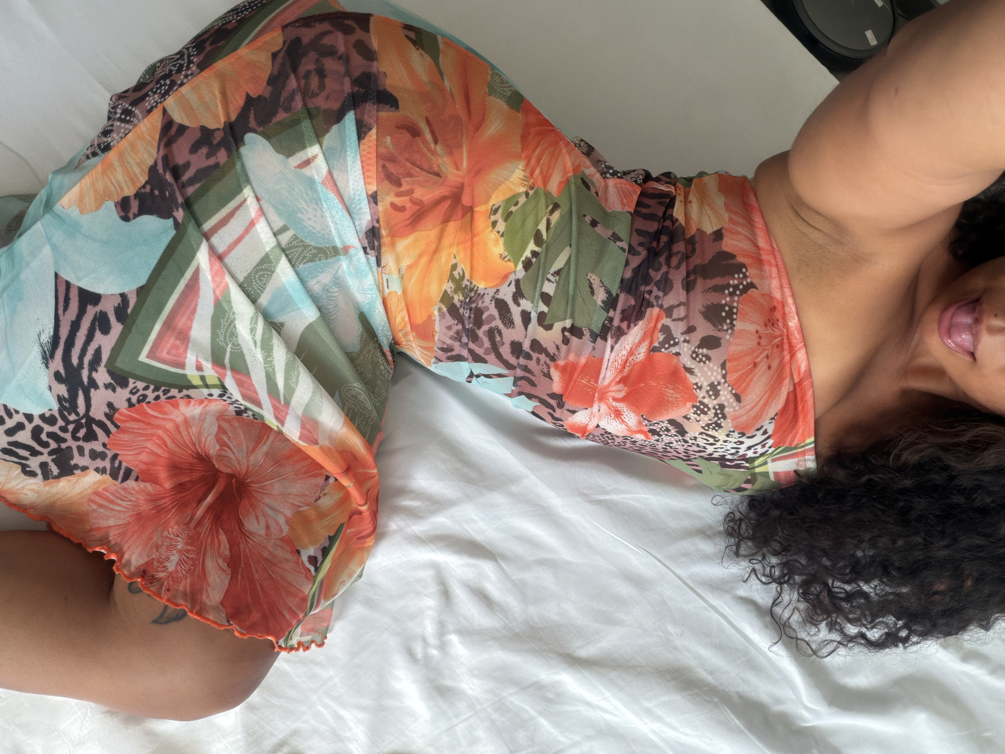 A woman lies on a white sheet in a supine position, wearing a floral-patterned dress. Her hair is dark and curly, and her right arm is raised above her head. The background is out of focus. There is no evidence to suggest the location is Barcelona, Spain.